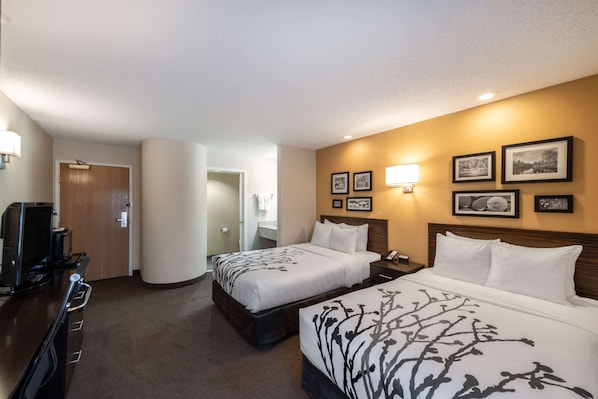 Habitación de hotel con dos camas, un escritorio con una computadora y un televisor. Hay cuadros en la pared y una lámpara sobre el escritorio.