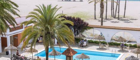 A pool area with lounge chairs and umbrellas, a beach in the background, and palm trees.