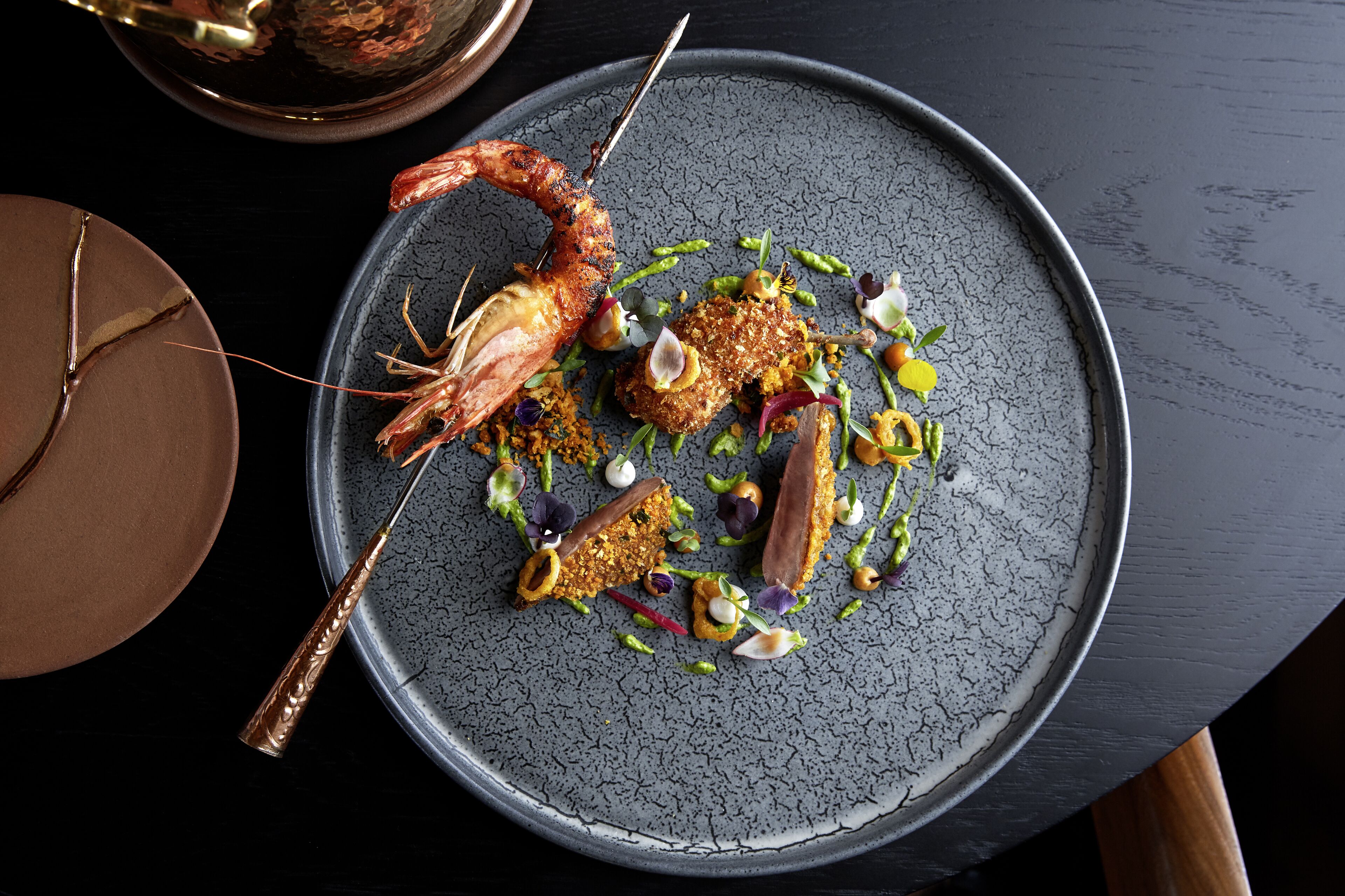 A plate of food with a variety of items including a grilled shrimp, a piece of bread, and some garnishes.