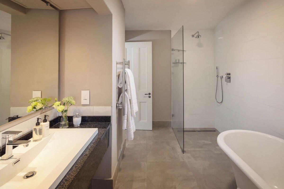 A modern bathroom with a large white bathtub, a glass-enclosed shower, a vanity with a mirror, and a door leading to another room.