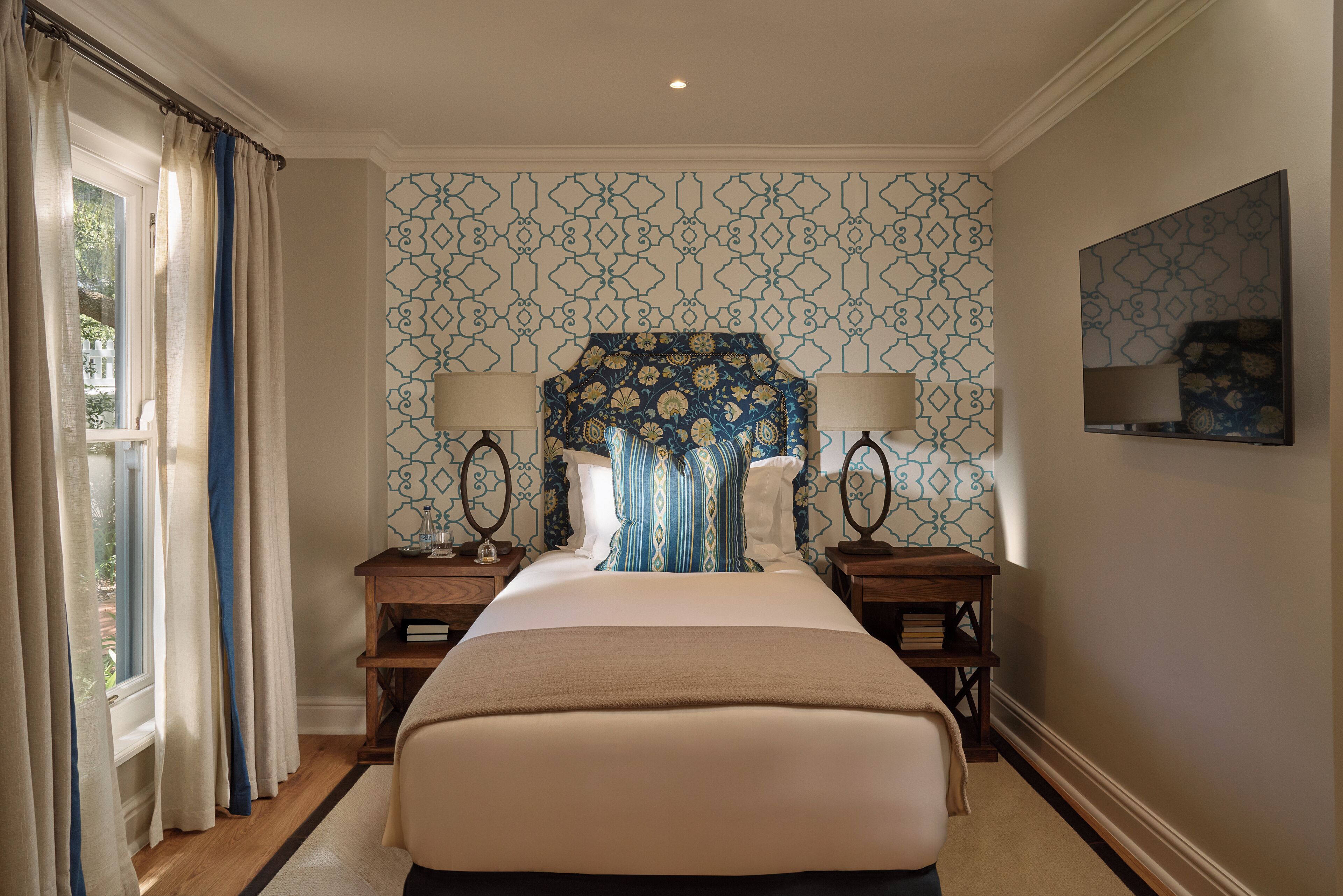 A hotel room with a large bed, two bedside tables, a nightstand, a window with curtains, patterned wallpaper, and a painting on the wall.