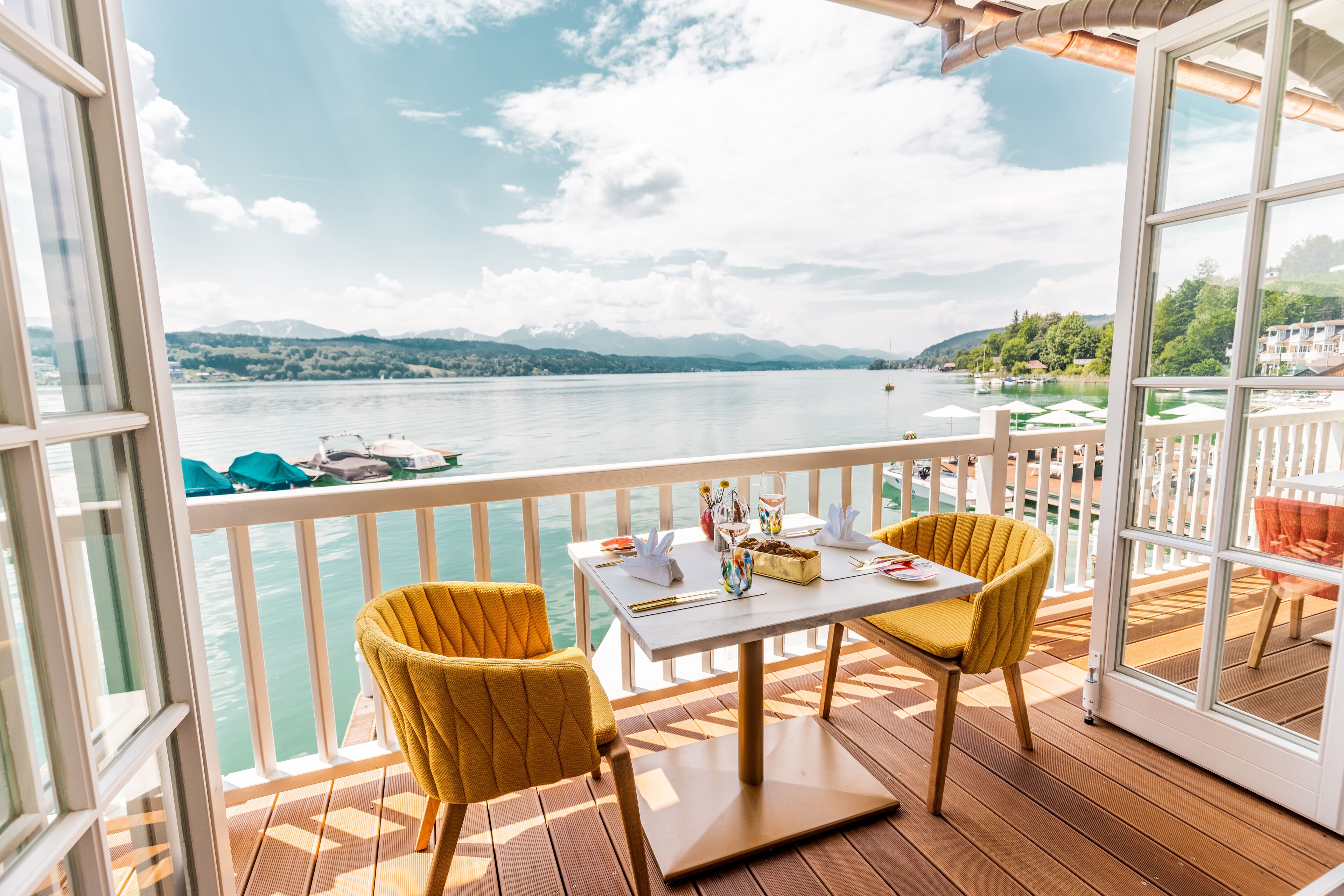 A balcony with a table set for two, overlooking a calm lake with boats and mountains in the distance.