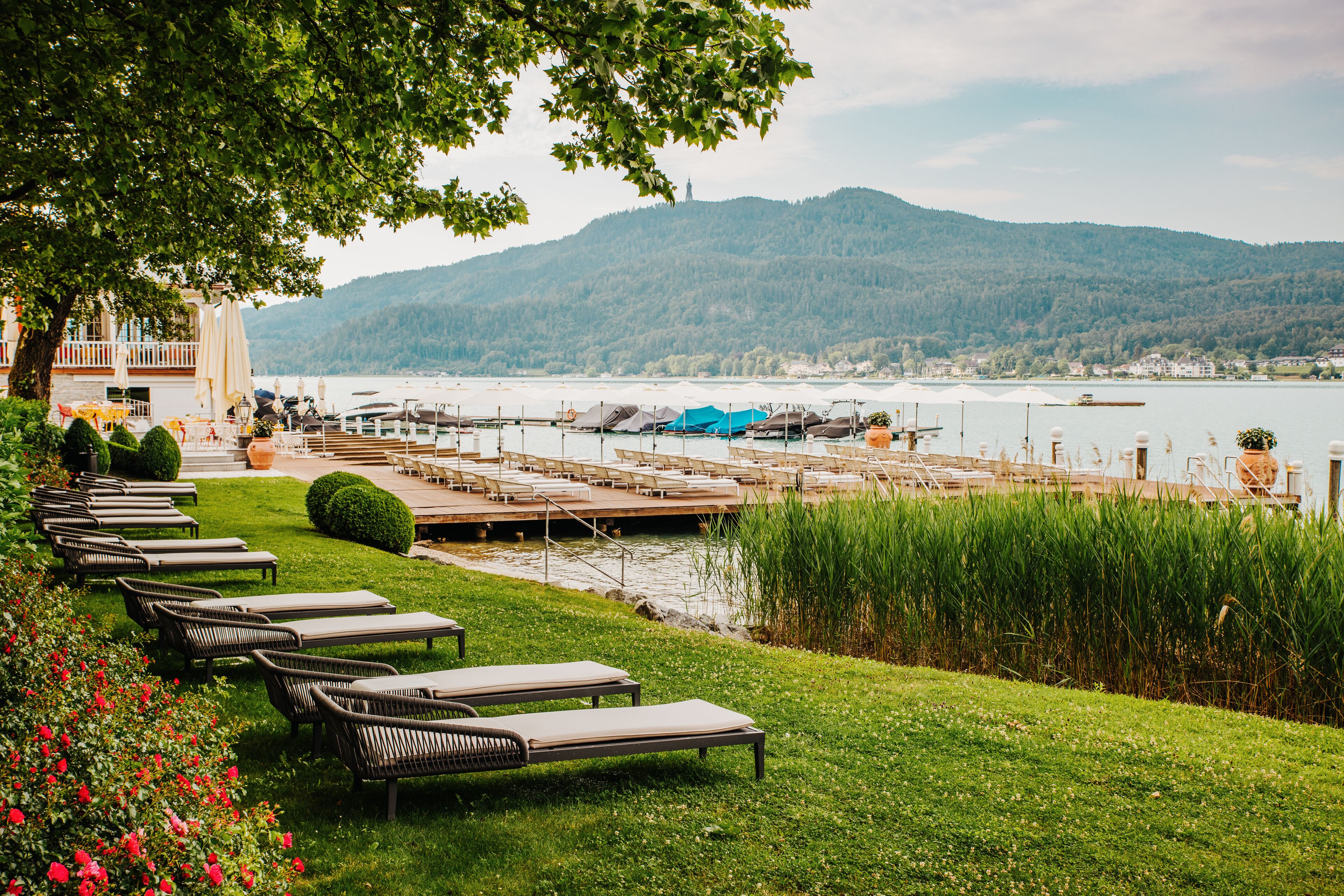 Ein Hotel am Seeufer mit Liegestühlen, einem Steg und Blick auf See und Berge.