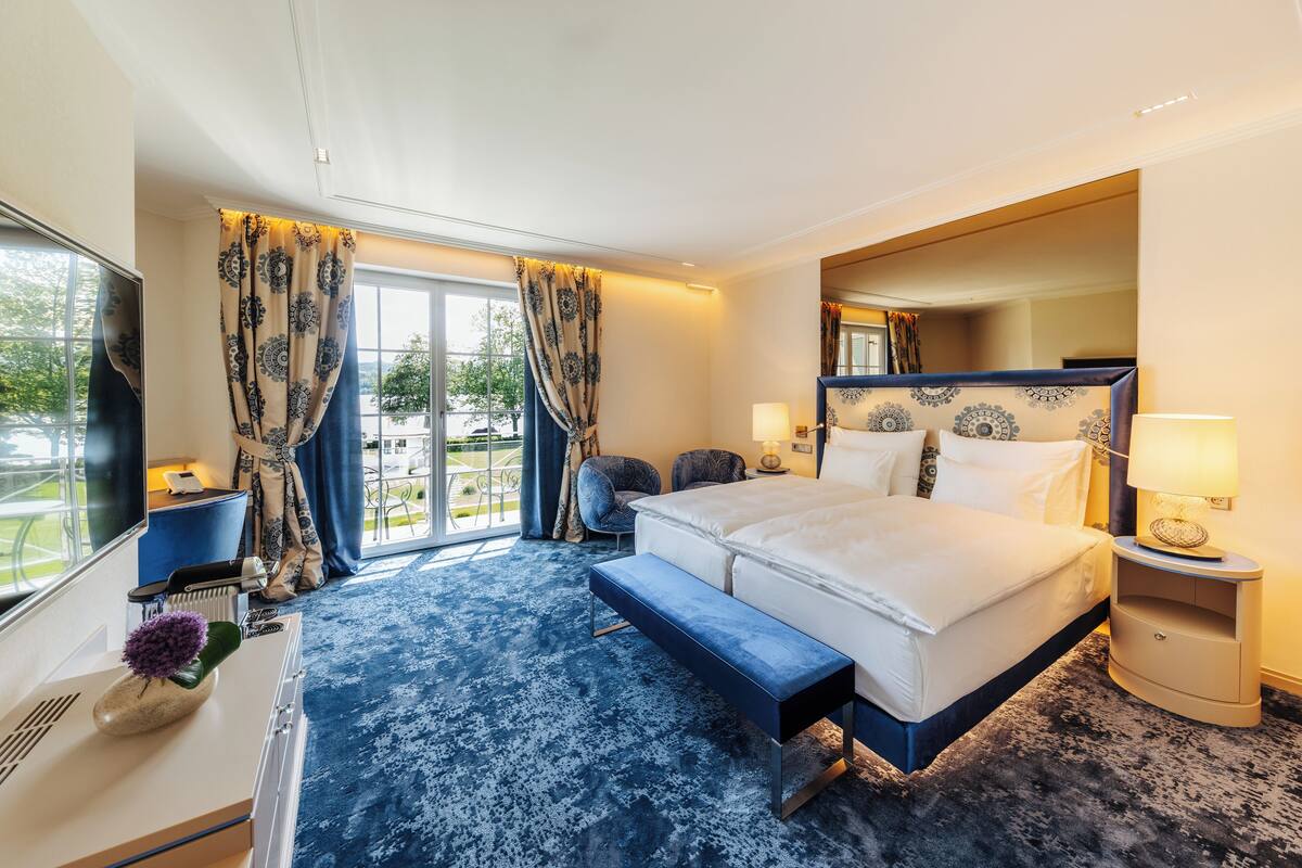 Habitación de hotel con una cama grande, un banco, una mesita de noche, un televisor y vistas al exterior.