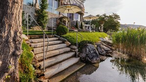 Una escalera de piedra que conduce a un edificio con fachada de vidrio, mesas al aire libre con sombrillas y un estanque rodeado de vegetación.