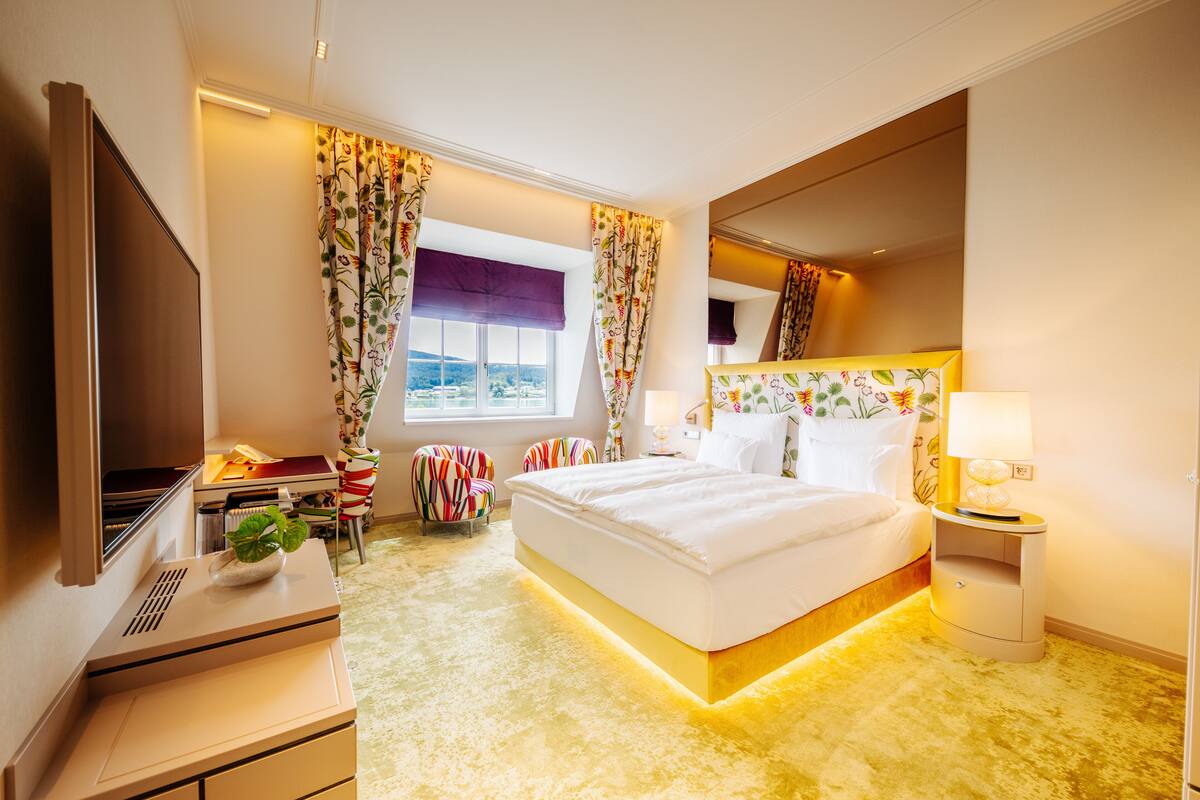 Habitación de hotel con cama, mesita de noche, lámpara y un amplio ventanal con cortinas.