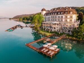 A large, multi-story hotel with a waterfront view, a dock with lounge chairs, and a pool area. - Hotel Schloss Seefels (Techelsberg am Worther See)