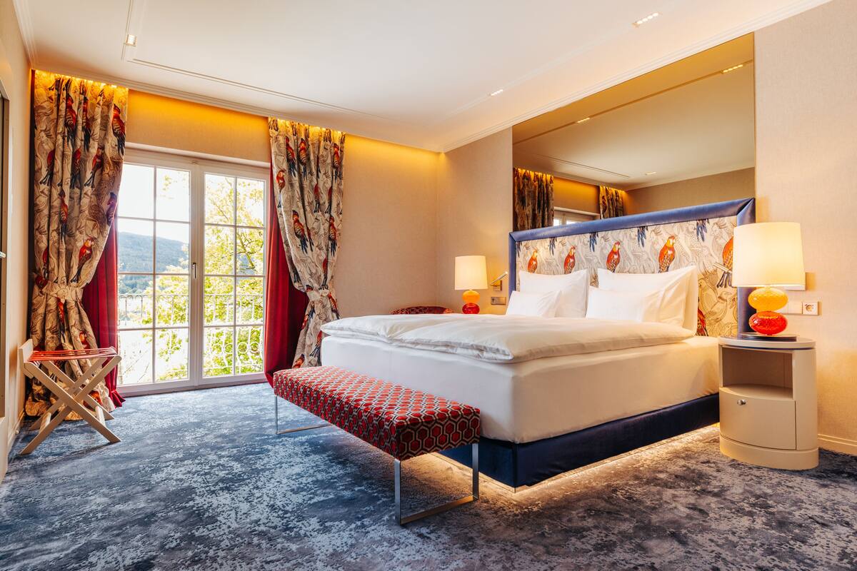 Una habitación de hotel moderna con una cama grande, un banco, una mesita de noche con lámparas y vistas al exterior a través de un ventanal.