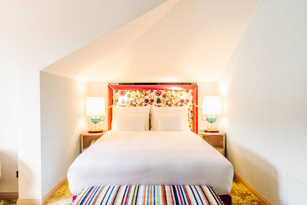 Una habitación de hotel con una cama grande, dos mesitas de noche con lámparas, un cabecero colorido y una alfombra a rayas.