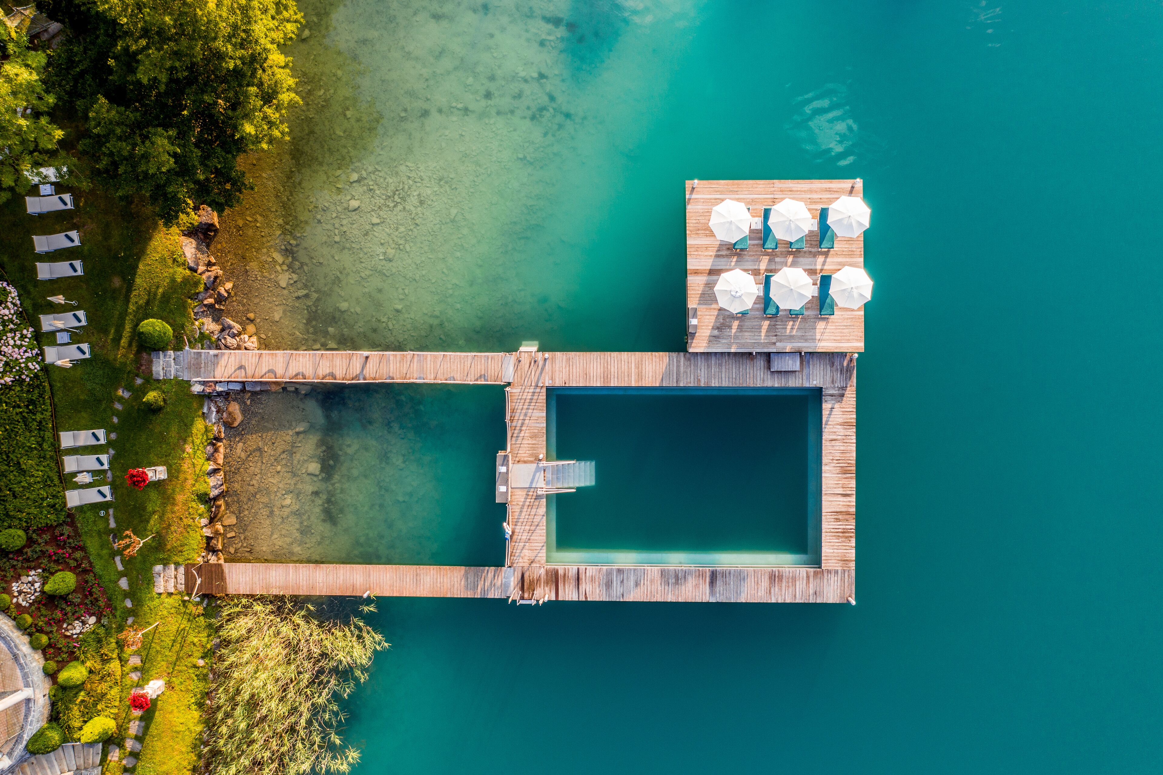 An aerial view of a wooden dock extending into a clear blue lake, with a pool and lounge area on the dock.
