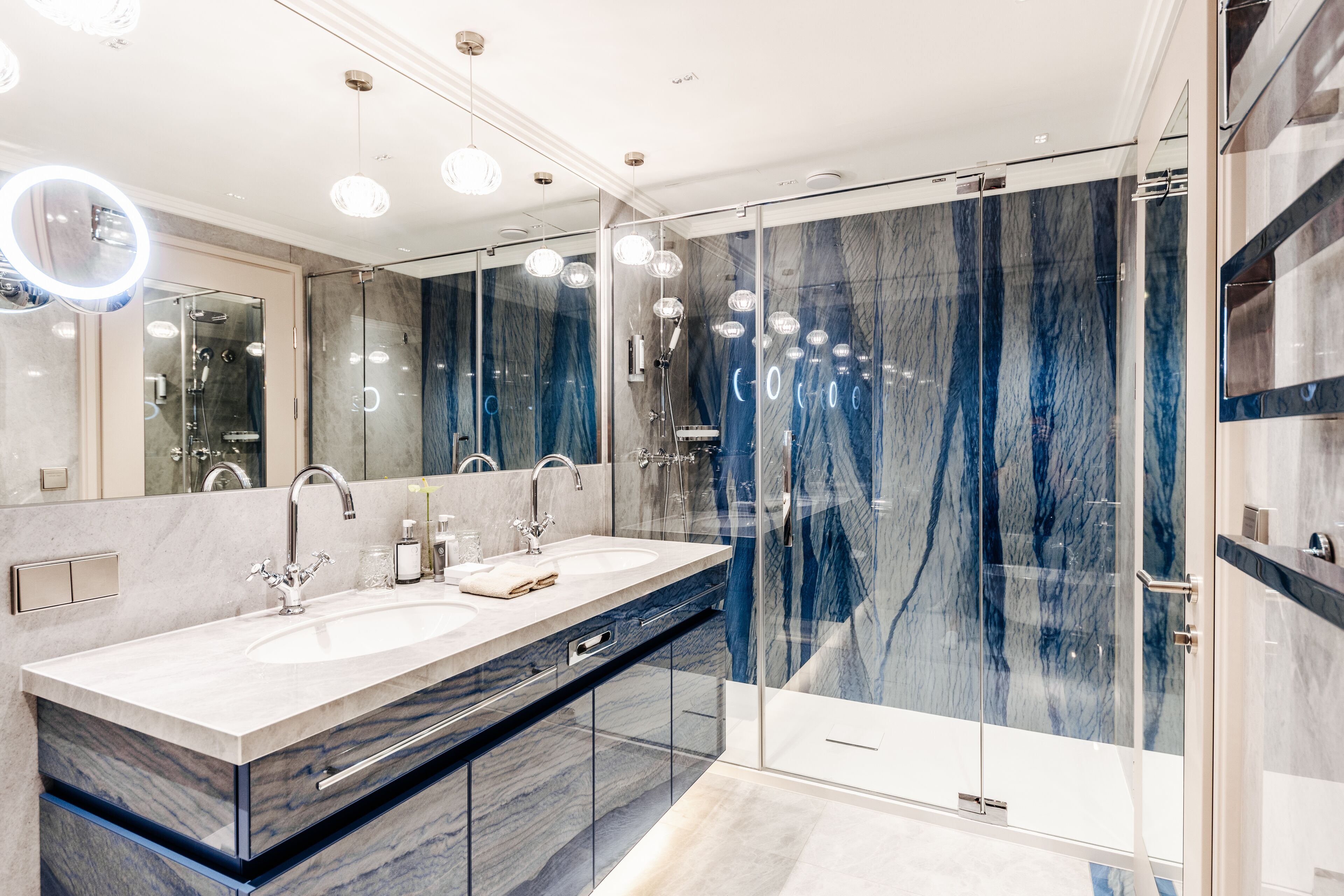 Un baño moderno con lavamanos doble, cabina de ducha con mampara de vidrio y paredes de mármol.