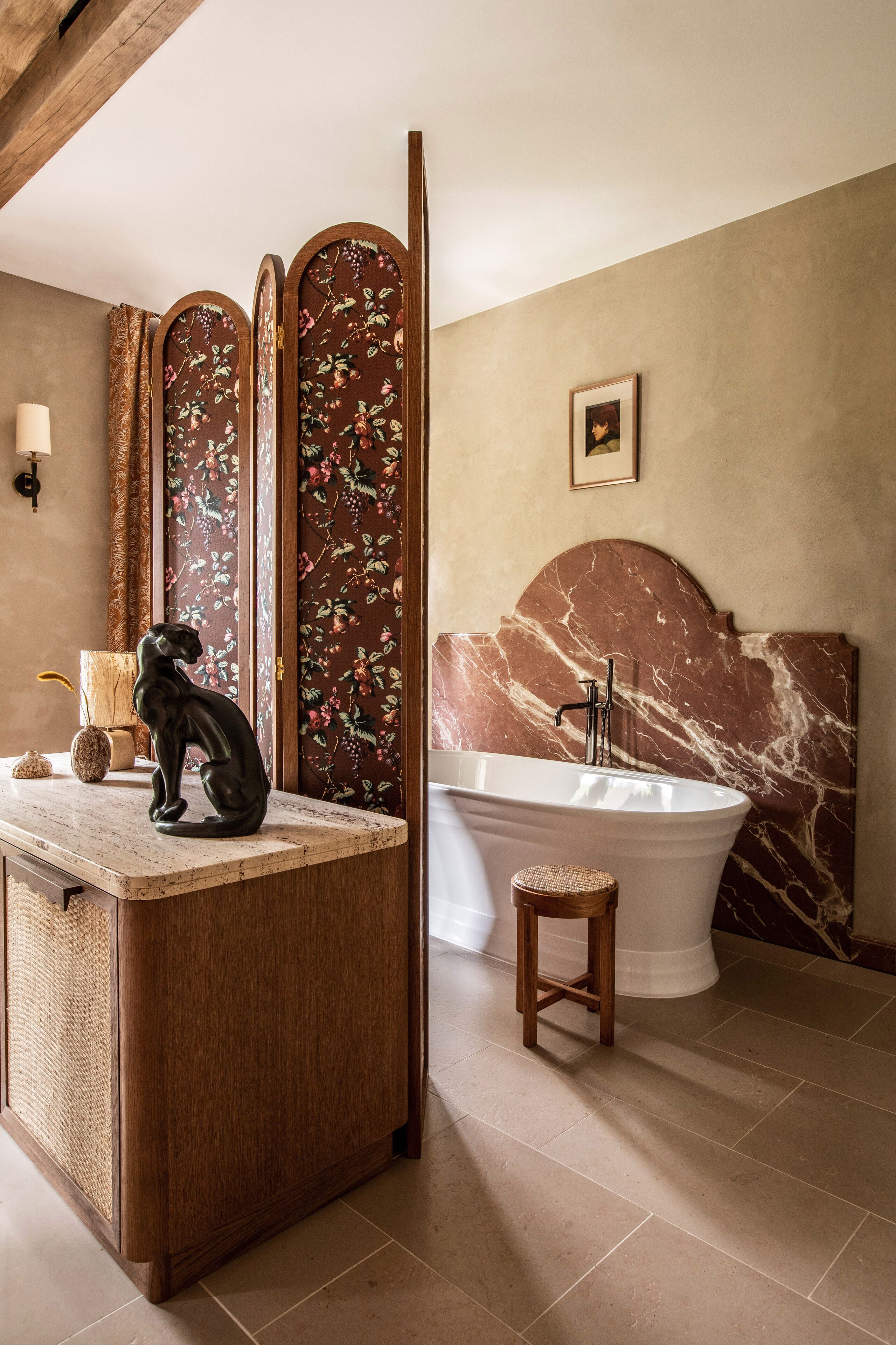 Une salle de bain avec une tête de lit en marbre, une baignoire sur pieds, un meuble-lavabo en bois orné d’une sculpture de chat noir et un paravent décoratif aux motifs floraux.