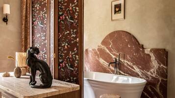 Une salle de bain avec une tête de lit en marbre, une baignoire sur pieds, un meuble-lavabo en bois orné d’une sculpture de chat noir et un paravent décoratif aux motifs floraux.