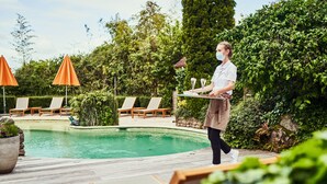 Zona de piscina con sillones de descanso y sombrillas, una terraza de madera y abundante vegetación al fondo.
