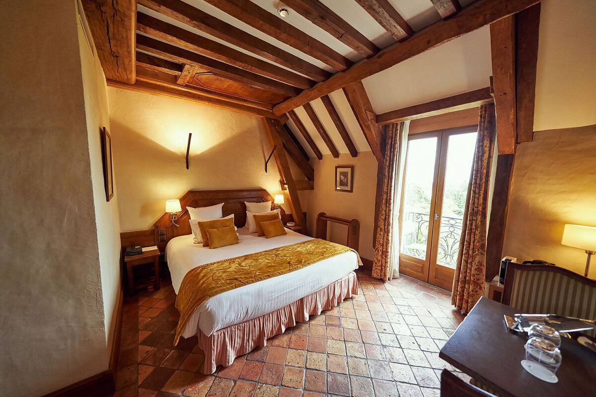 Un dormitorio con una cama grande, vigas de madera, suelo de baldosas y vistas a través de una ventana.