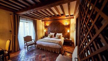 Habitación de hotel con una cama grande, cabecero de madera y suelo de cuadros. Hay dos sillas y una mesa con tazas y una cuchara.