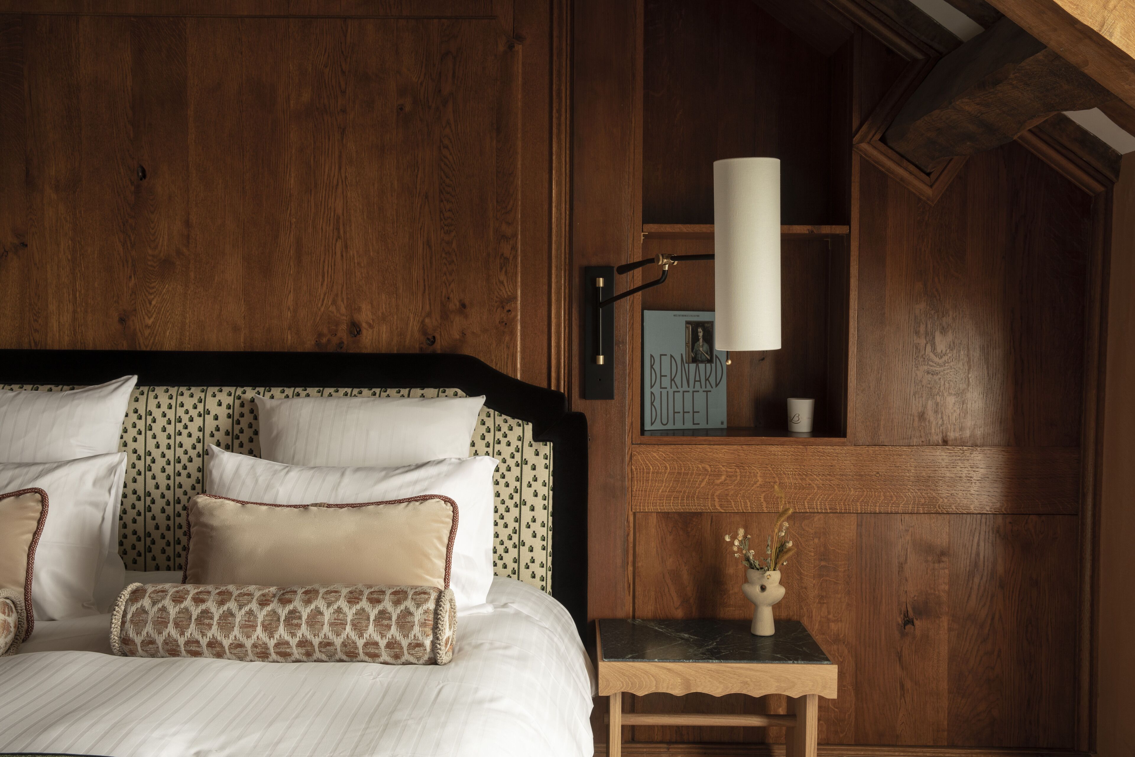 Un dormitorio de madera con una cama, almohadas, una mesita de noche, una lámpara y un cuadro en la pared.