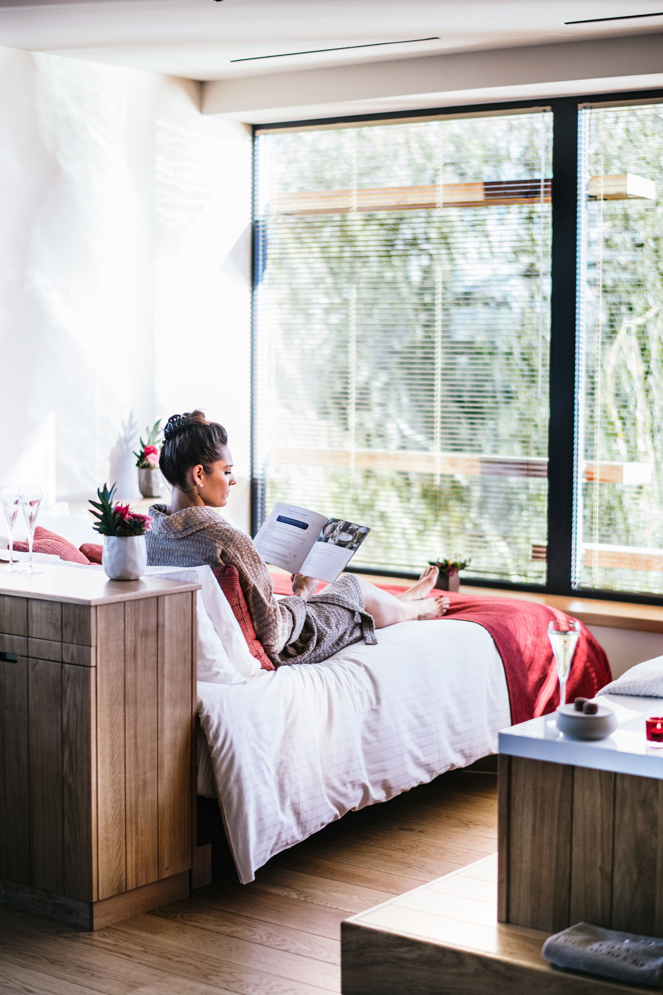 A modern bedroom with a large window, a wooden bedside table, a bed with white bedding, and a small plant on the table.