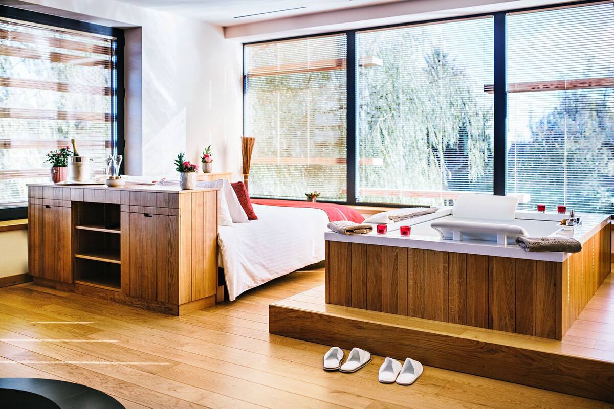 Una habitación de hotel moderna con piso de madera, una cama con ropa blanca, un escritorio de madera y ventanas grandes con persianas.