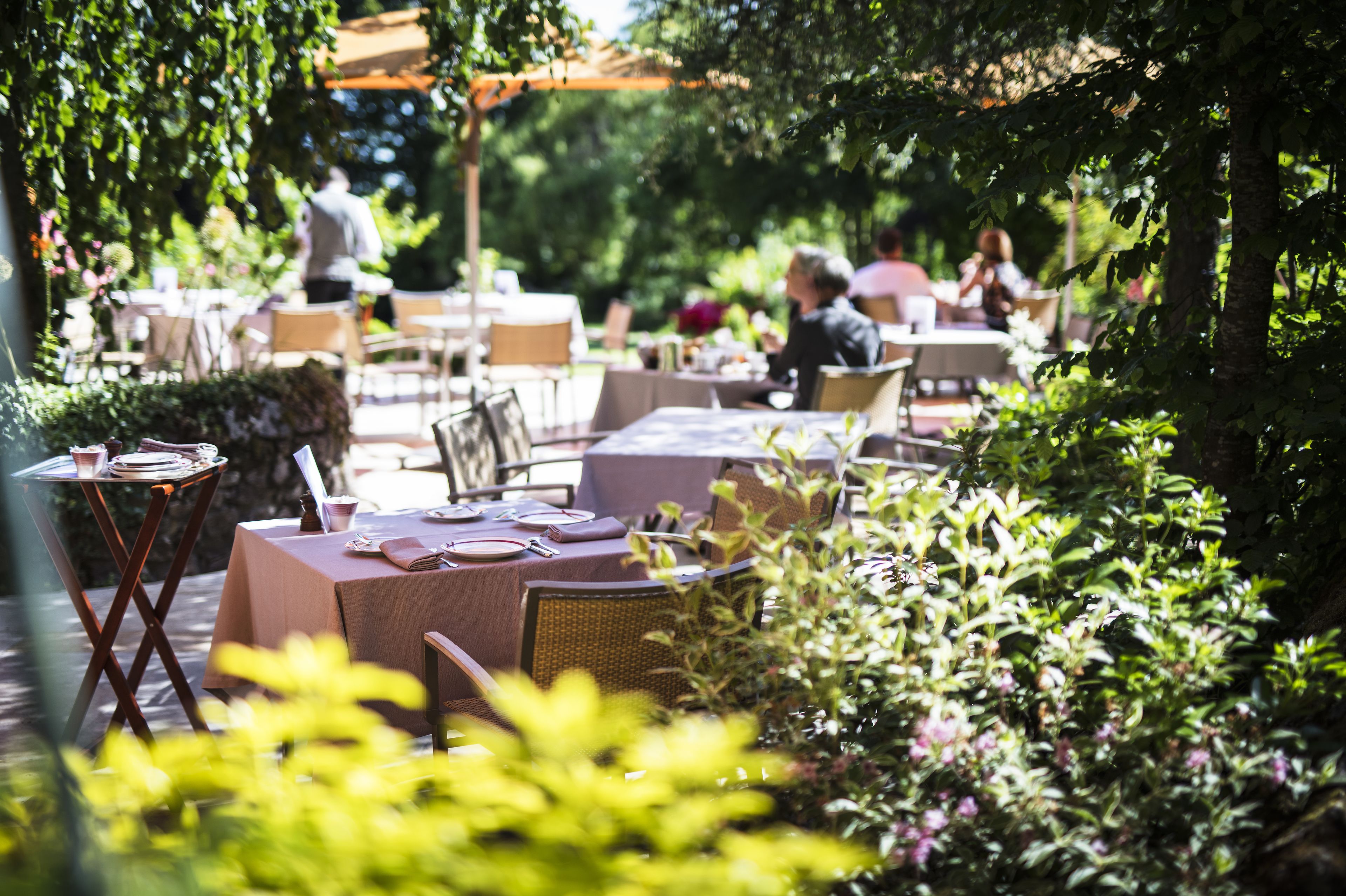 An outdoor dining area with tables and chairs, surrounded by greenery.