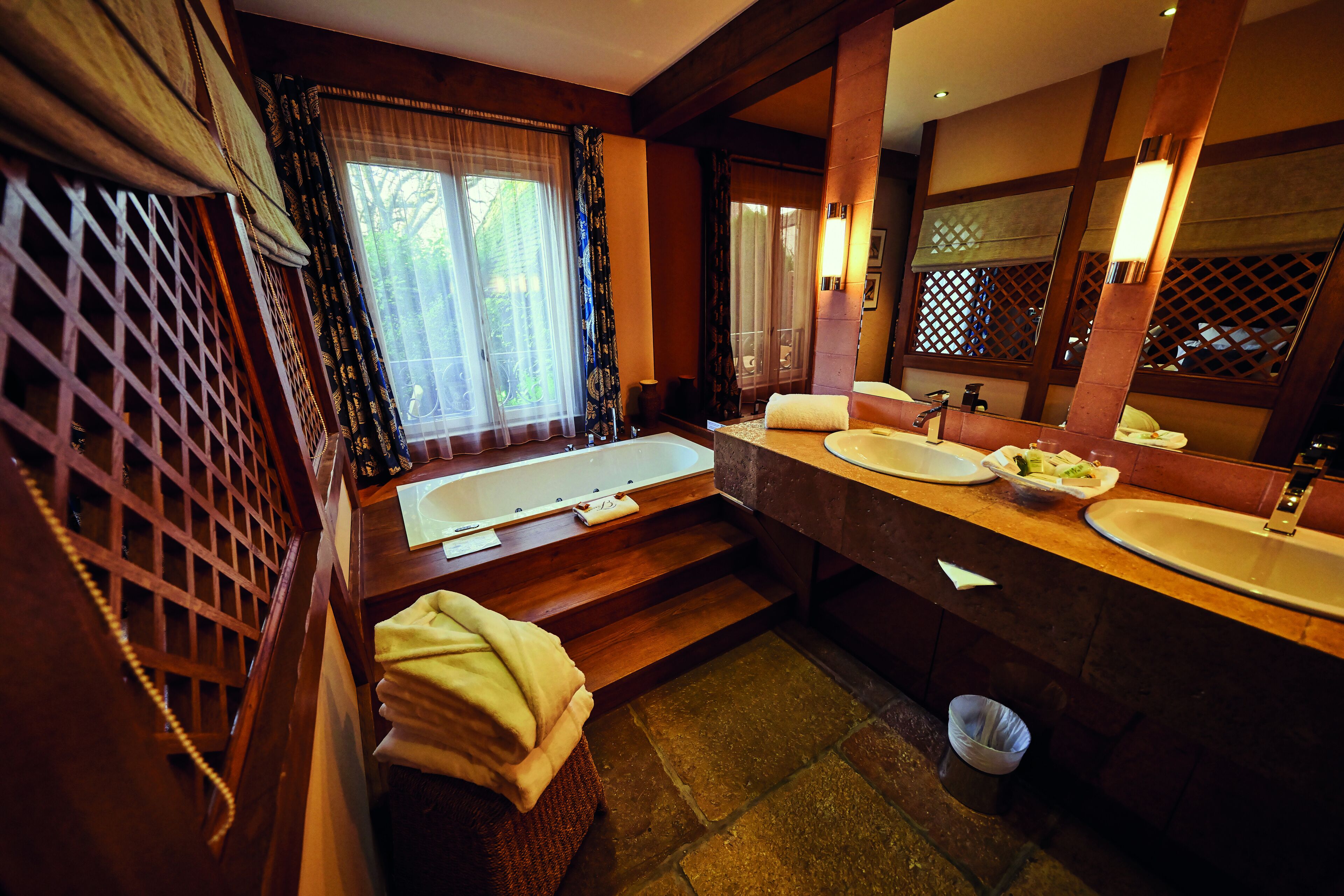 Une salle de bain avec une grande baignoire, deux lavabos et un miroir. Des marches en bois mènent à la baignoire, et une serviette est posée sur un banc.