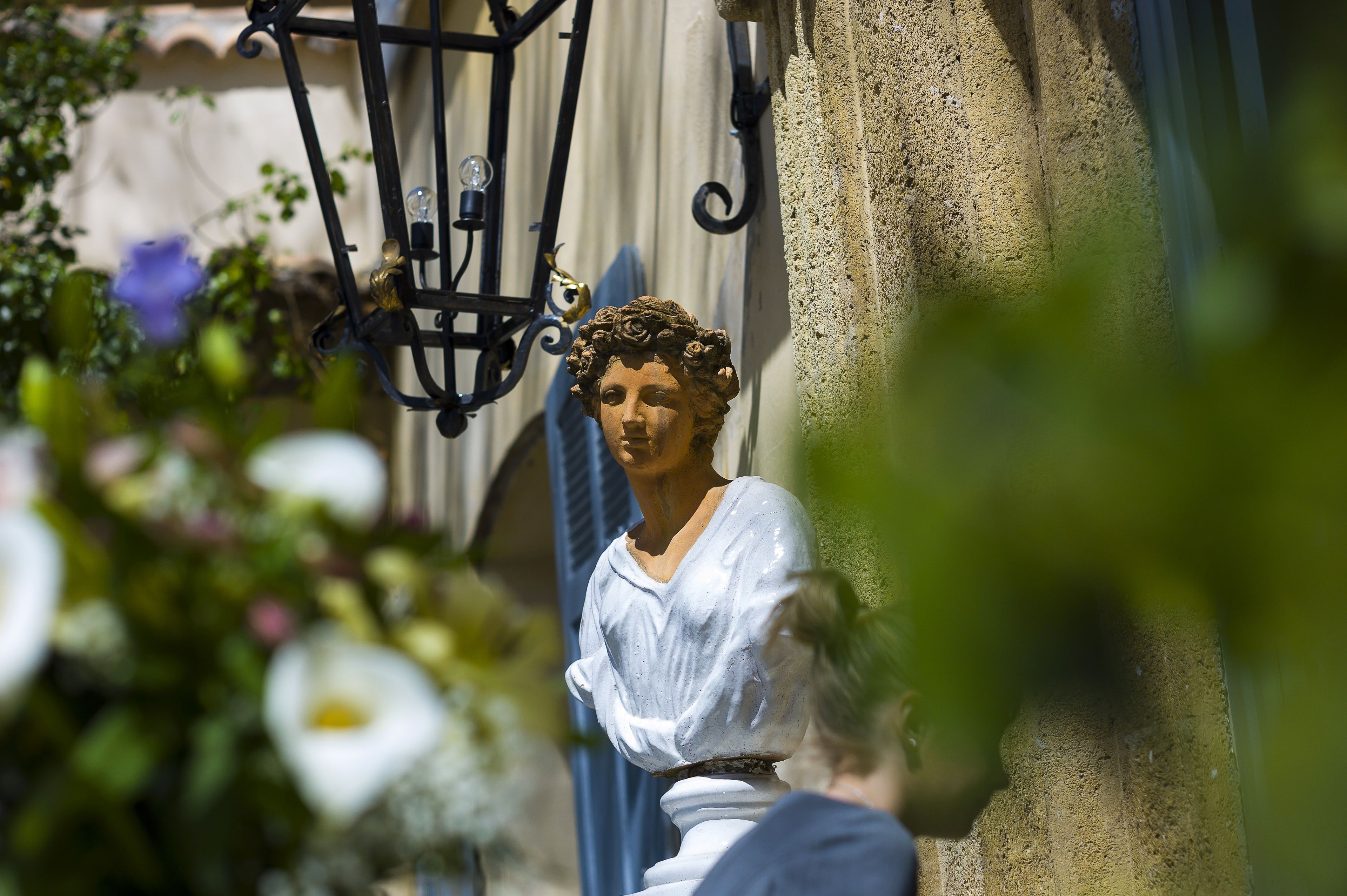 Eine Statue einer Frau in weißer Kleidung, neben einem Gebäude mit einer strukturierten Wand und einem schwarzen Metall-Laternenpfahl.