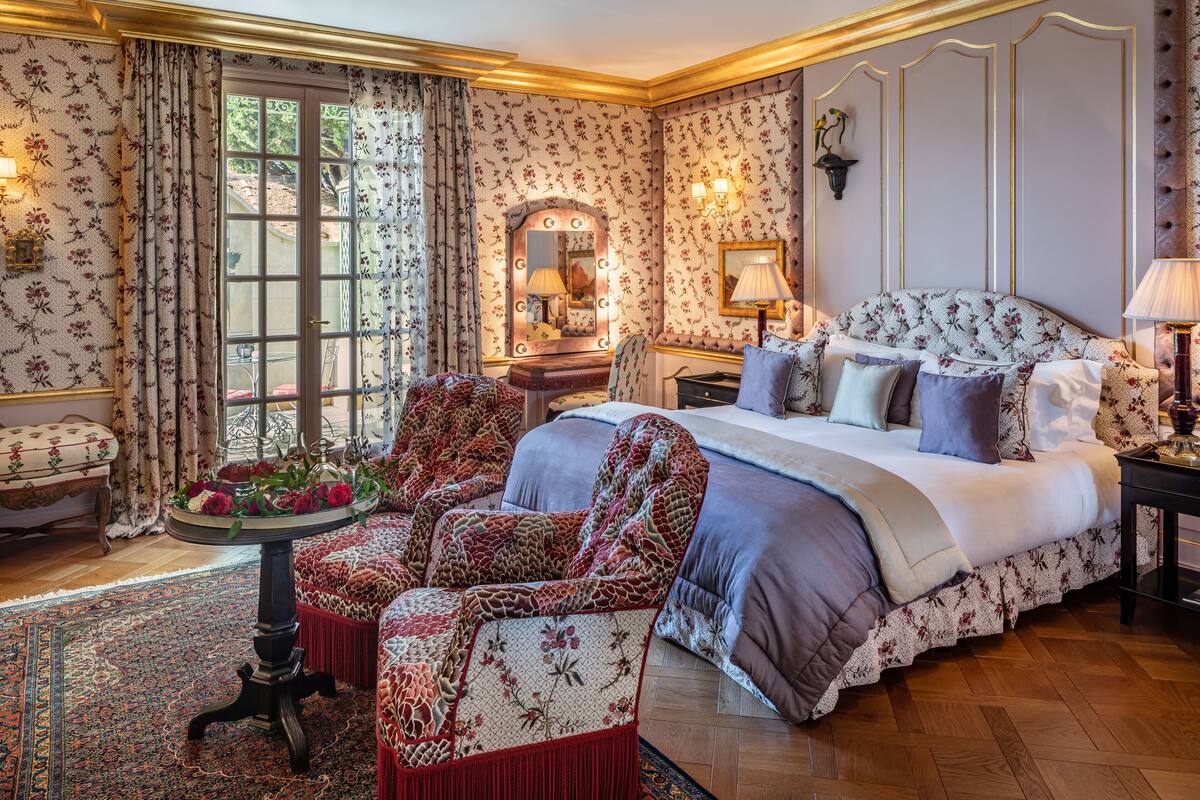 Une chambre à coucher avec un grand lit, un coin salon avec un fauteuil, une petite table agrémentée de fleurs et un miroir avec éclairage.