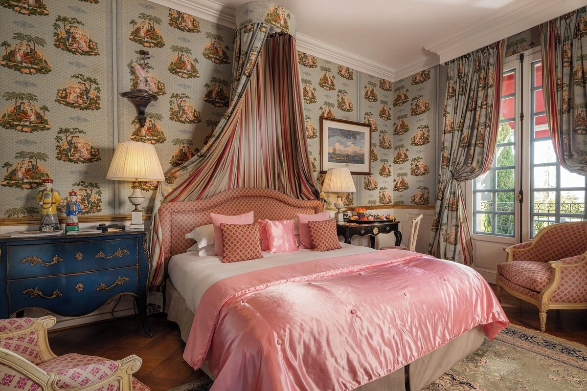 A bedroom with a canopy bed, patterned wallpaper, a blue dresser, and a chair.