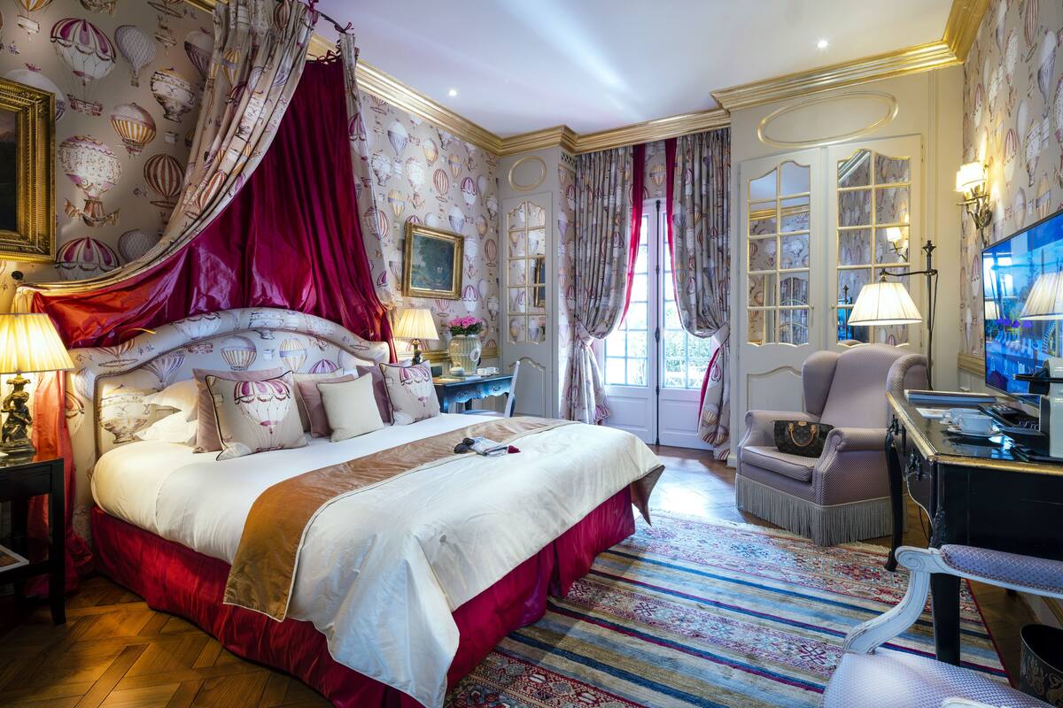 Une chambre spacieuse avec un grand lit, un bureau et une chaise. Il y a une fenêtre avec des rideaux et une vue sur l’extérieur.