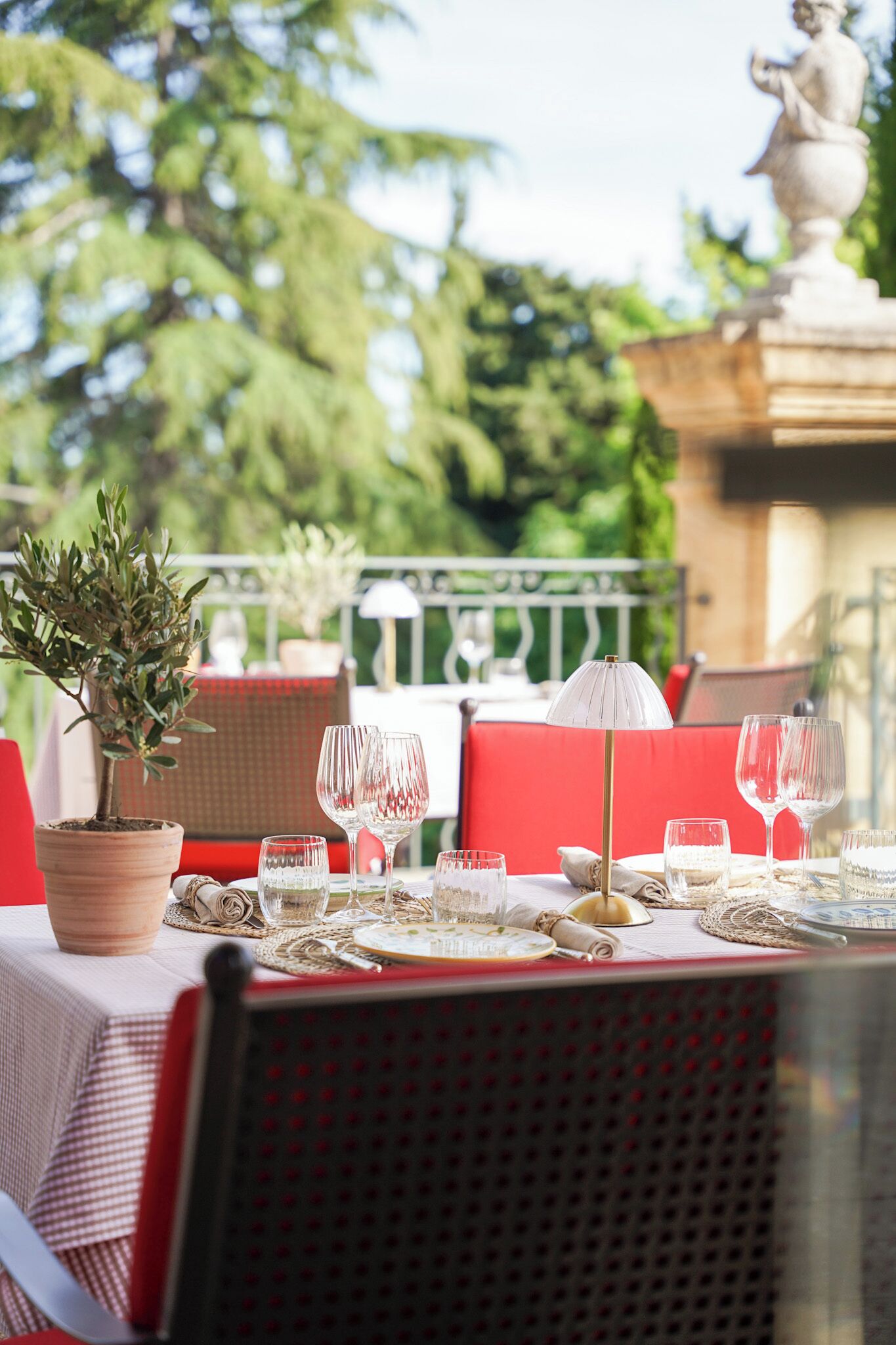 Un área de comedor al aire libre con una mesa preparada para dos personas, que incluye una planta en maceta, sillas con cojines rojos y un paraguas blanco.