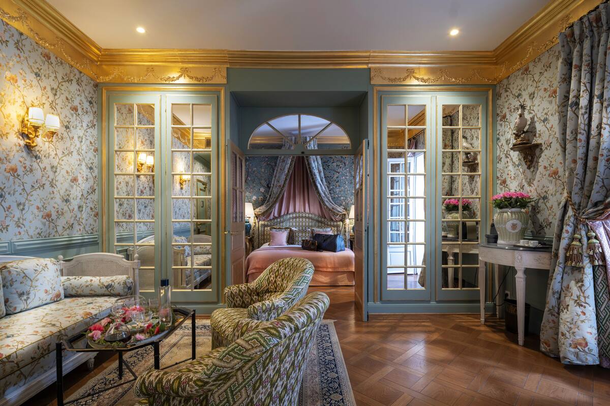Une chambre à coucher luxueuse, dotée d’un grand lit, d’un coin salon avec un canapé et des fauteuils, et offrant une vue sur un couloir agrémenté d’un miroir et d’étagères.