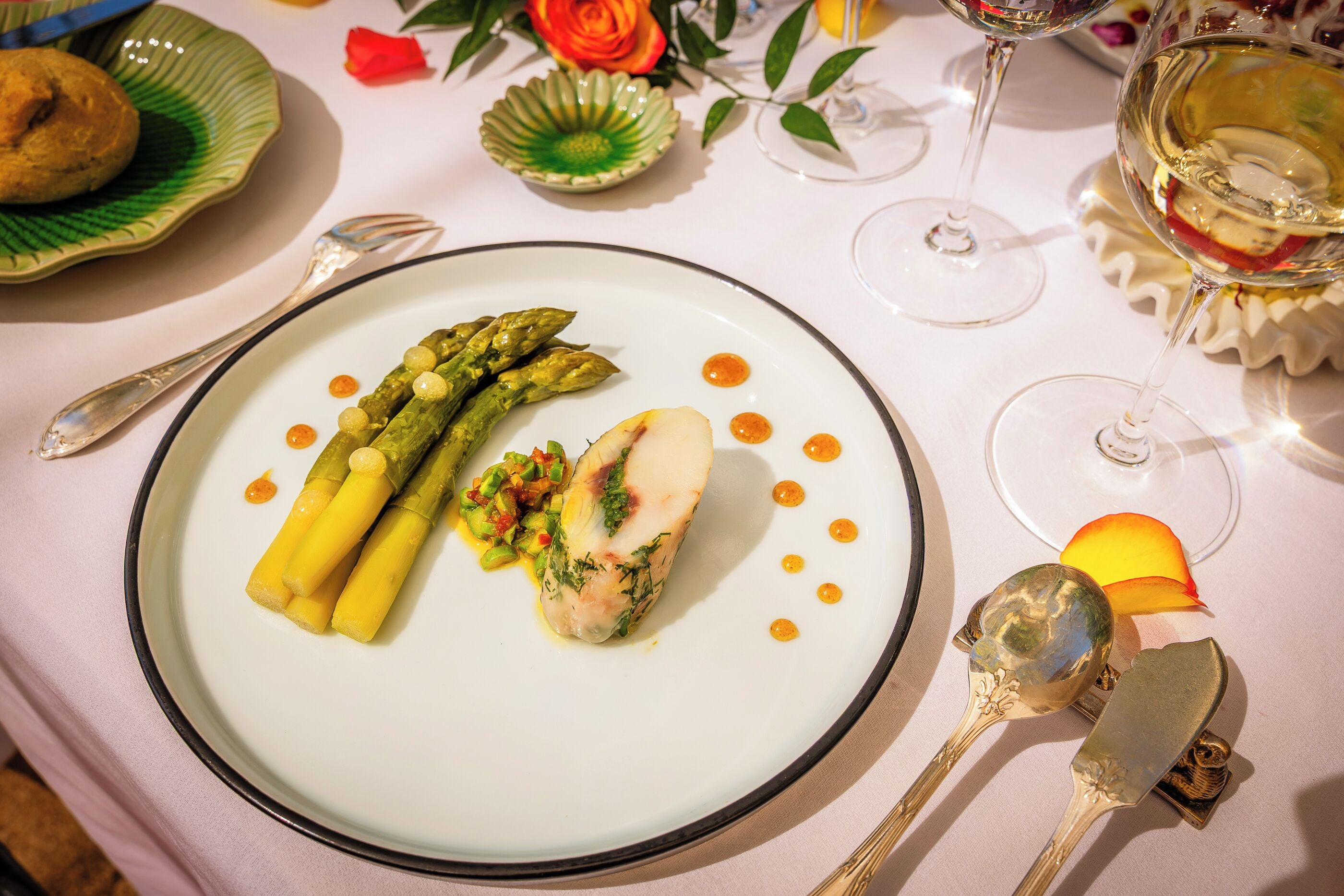 A plate of food with asparagus and a piece of fish, accompanied by a spoon and a fork on a table.