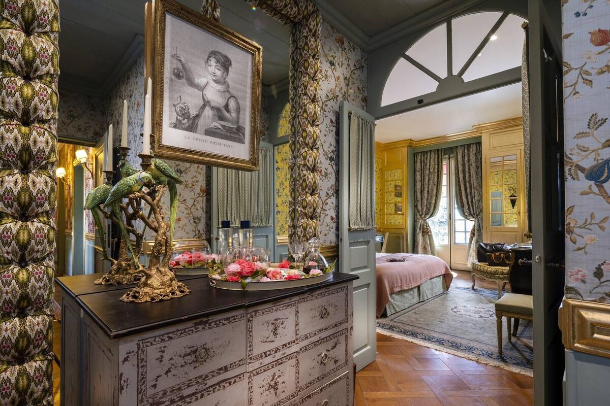 Une pièce avec une commode ancienne, un portrait encadré et un lit recouvert d’une couvre-lit rose.