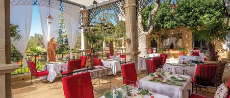 An outdoor restaurant with red chairs and white tablecloths, surrounded by greenery and statues.