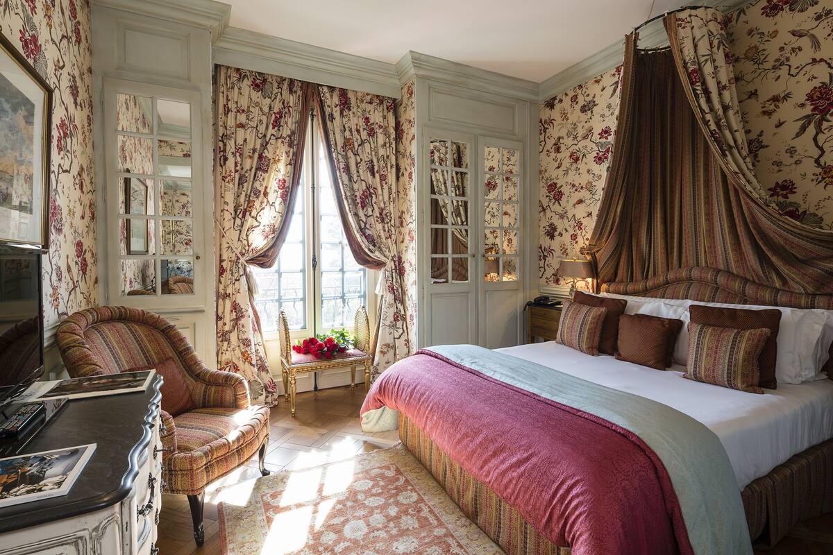 Une chambre à coucher classique, dotée d’un grand lit, d’un coin salon avec un fauteuil et d’un bureau avec une lampe.
