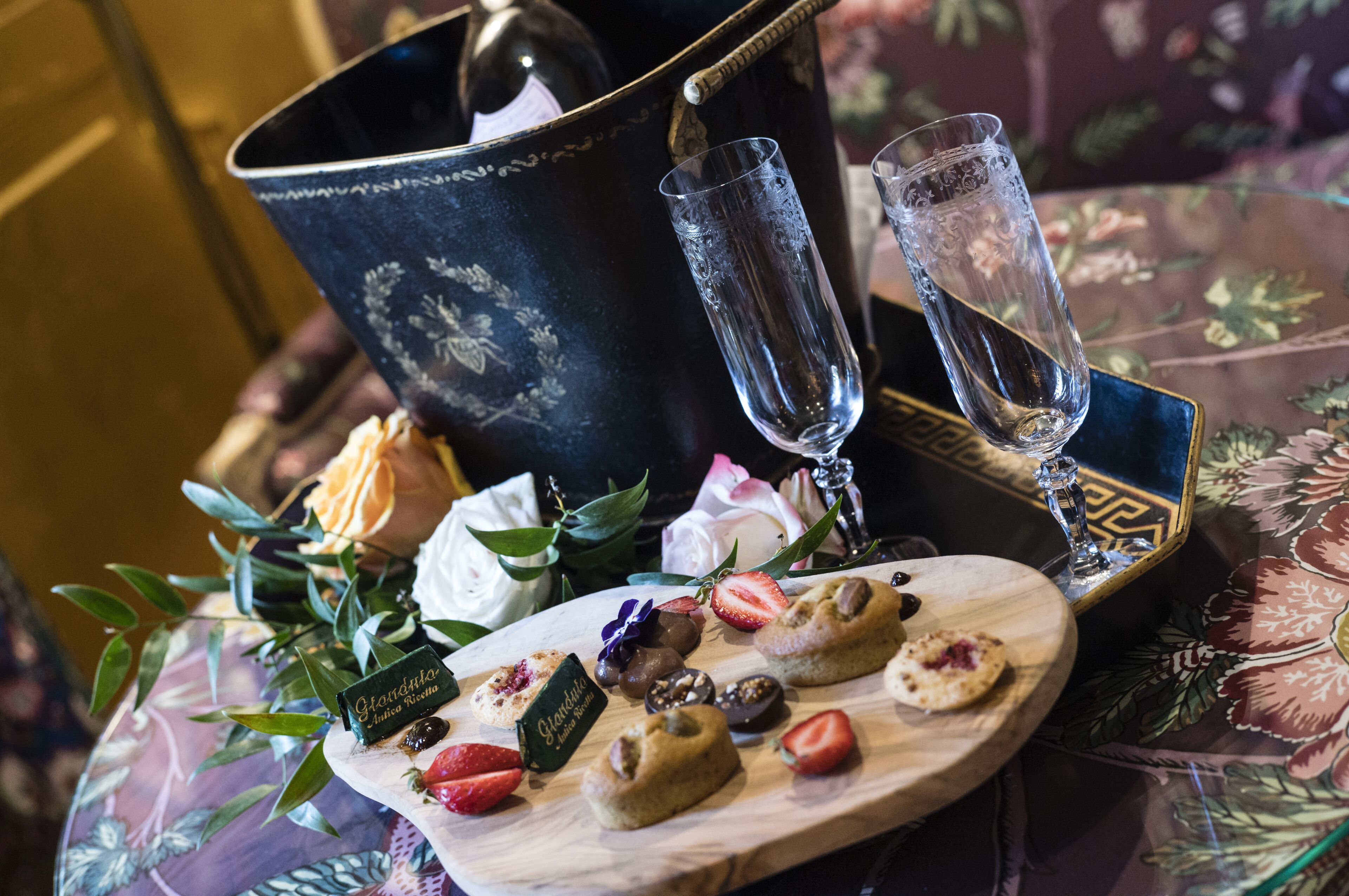 Une table recouverte d’une nappe à motifs floraux, une bouteille de champagne, deux verres, ainsi qu’un assortiment de pâtisseries et de chocolats.