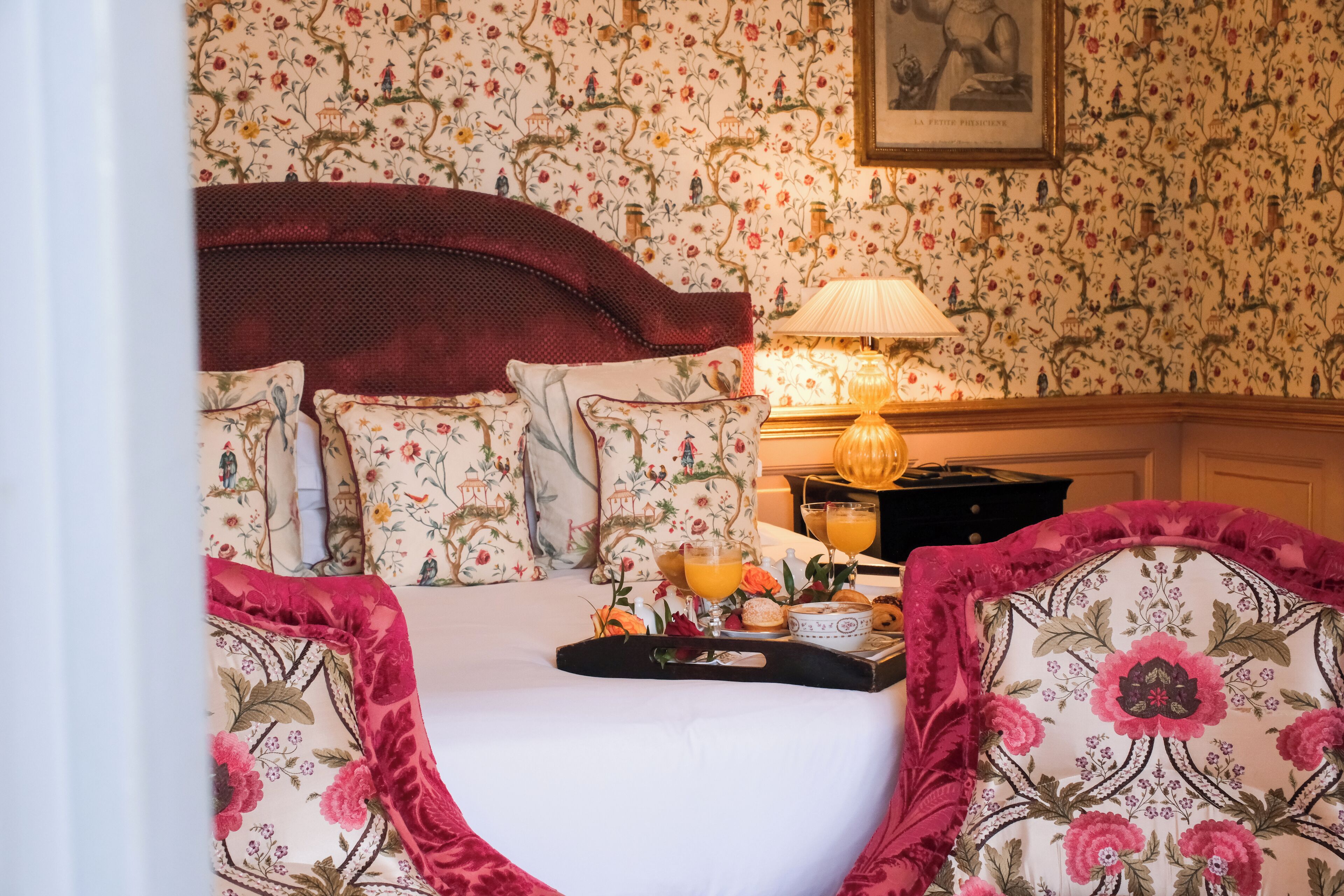 Ein ordentlich gemachtes Bett mit Blumenmuster auf der Bettwäsche, ein Nachttisch mit Lampe und ein Tablett mit Frühstücksgebäck.