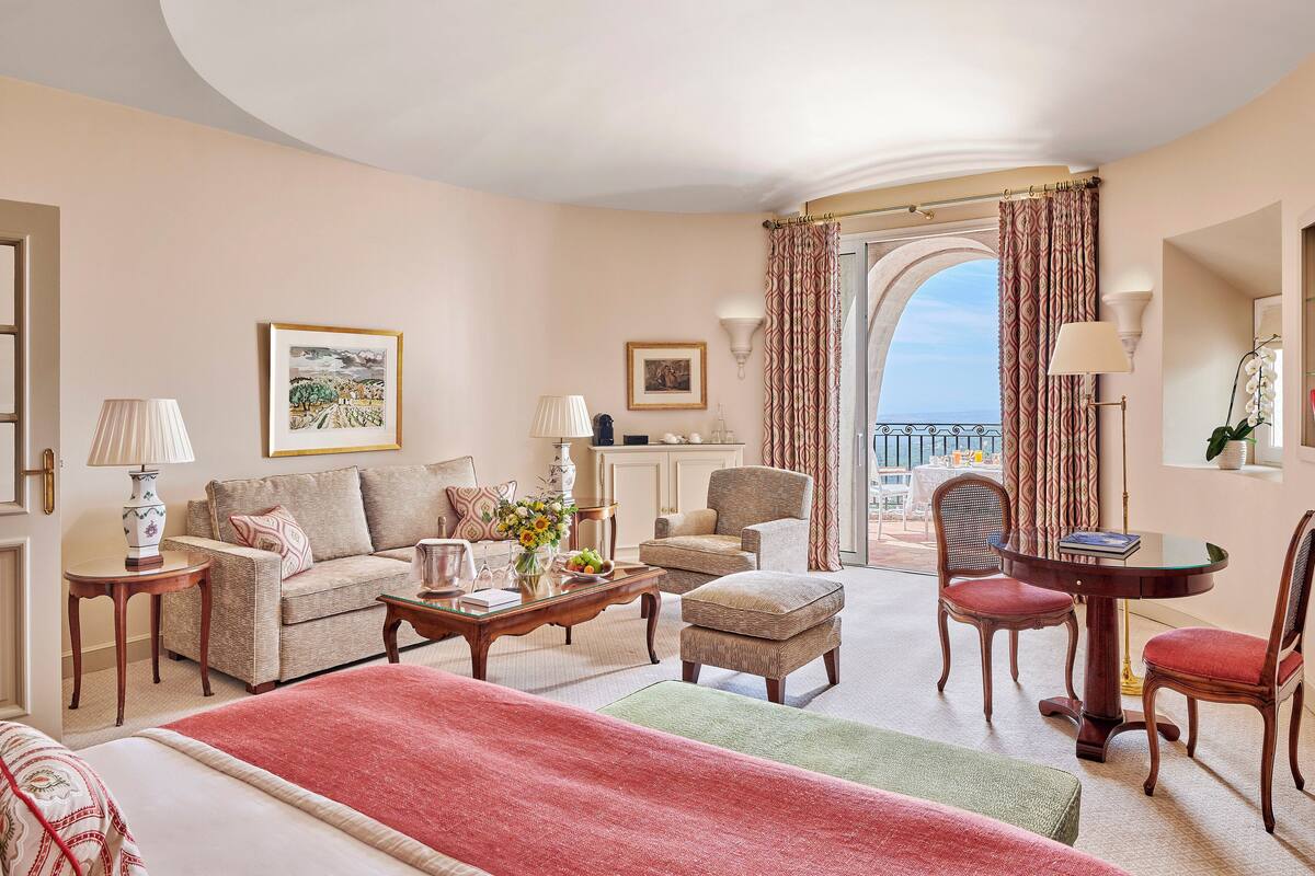 A hotel room with a bed, a sofa, a dining table with chairs, a small table with a lamp, and a view of the sea through an arched doorway.