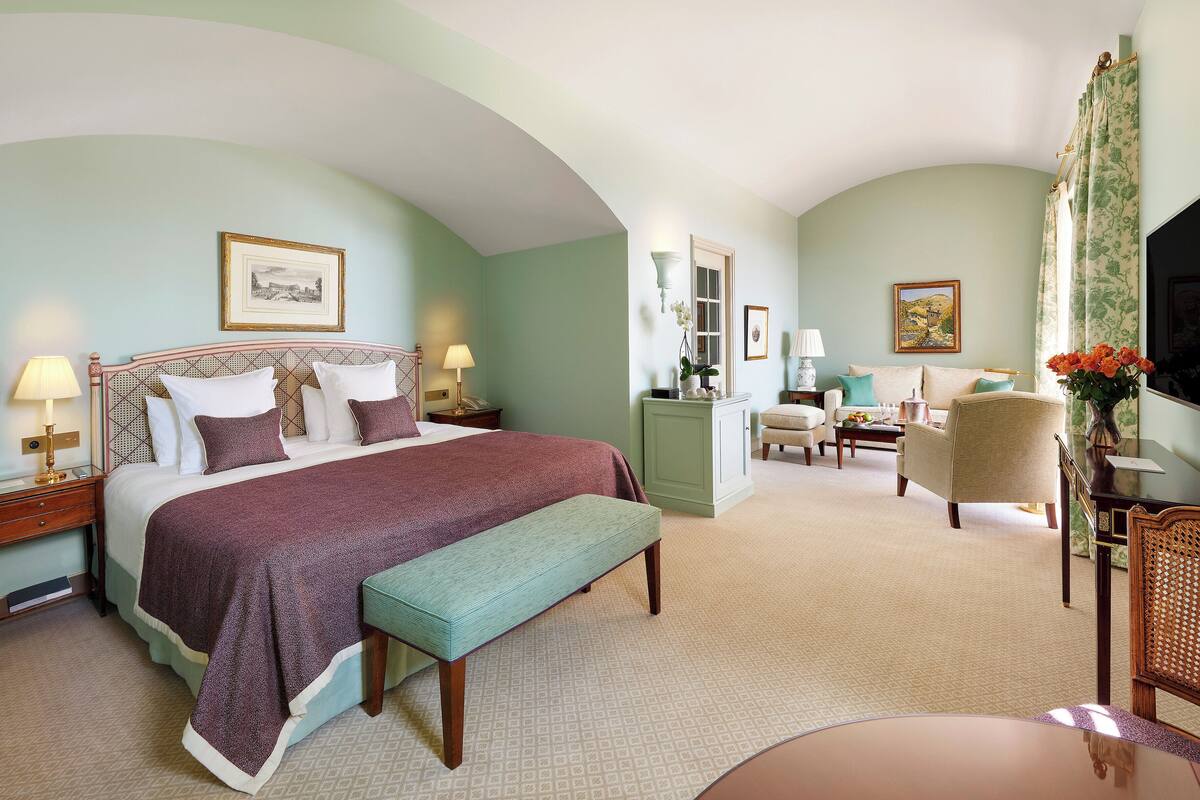 A spacious bedroom with a large bed, a sitting area with a chair and table, and a television.