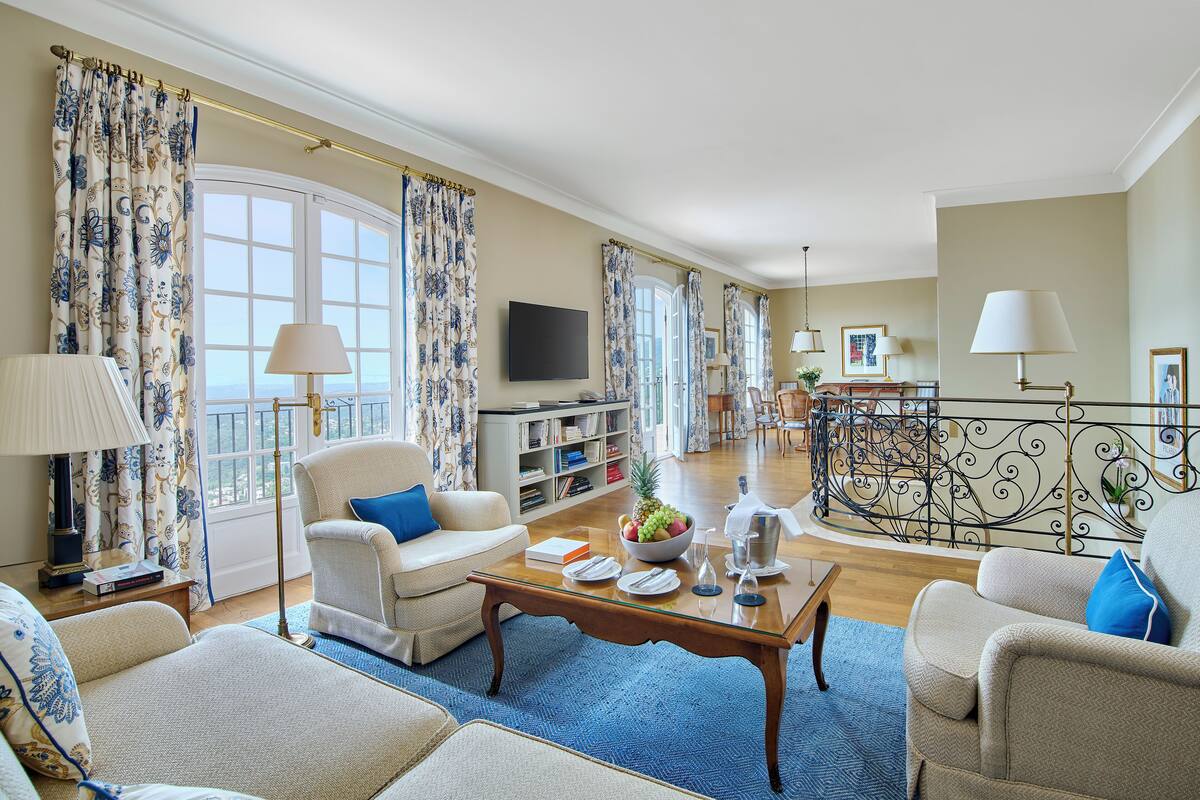 A spacious living room with a large window, a flat-screen TV, a coffee table, and a dining area with a fruit bowl.