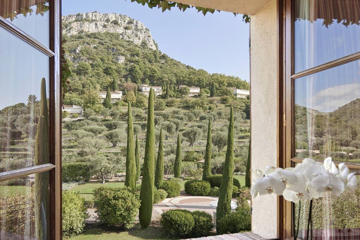 A view from a window overlooking a mountainous landscape with cypress trees and a well-maintained garden.
