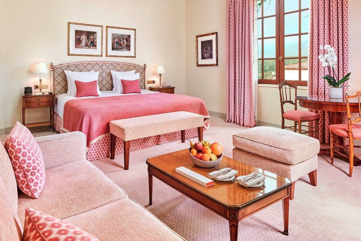 A hotel room with a large bed, two bedside tables, a sitting area with a sofa and armchairs, a small dining table with fruit and napkins, and a window with curtains.