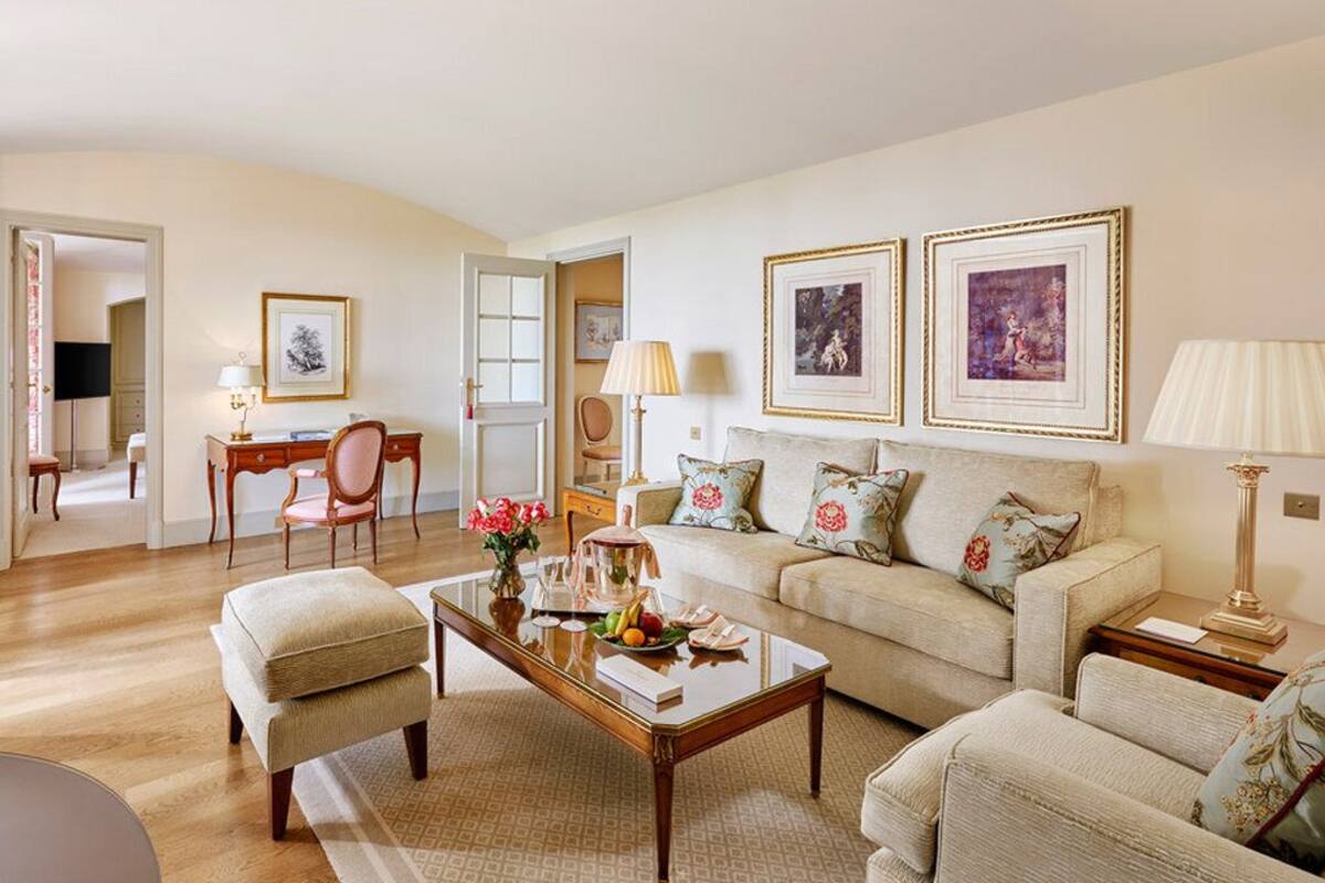 A living room with a sofa, armchairs, a coffee table, and a TV. There are framed pictures on the wall and a lamp on a side table.