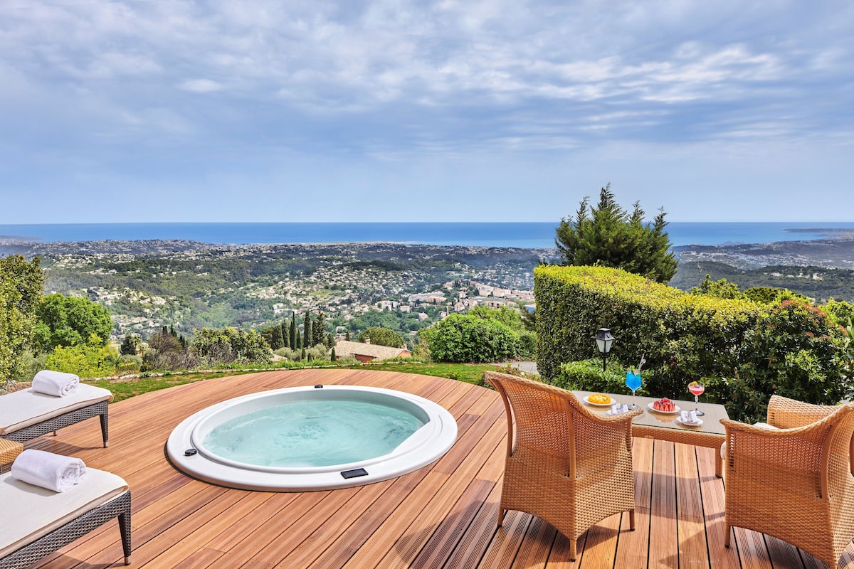 A wooden deck with a hot tub, lounge chairs, and a table with a drink. A scenic view of a coastal town and hills is visible in the background.