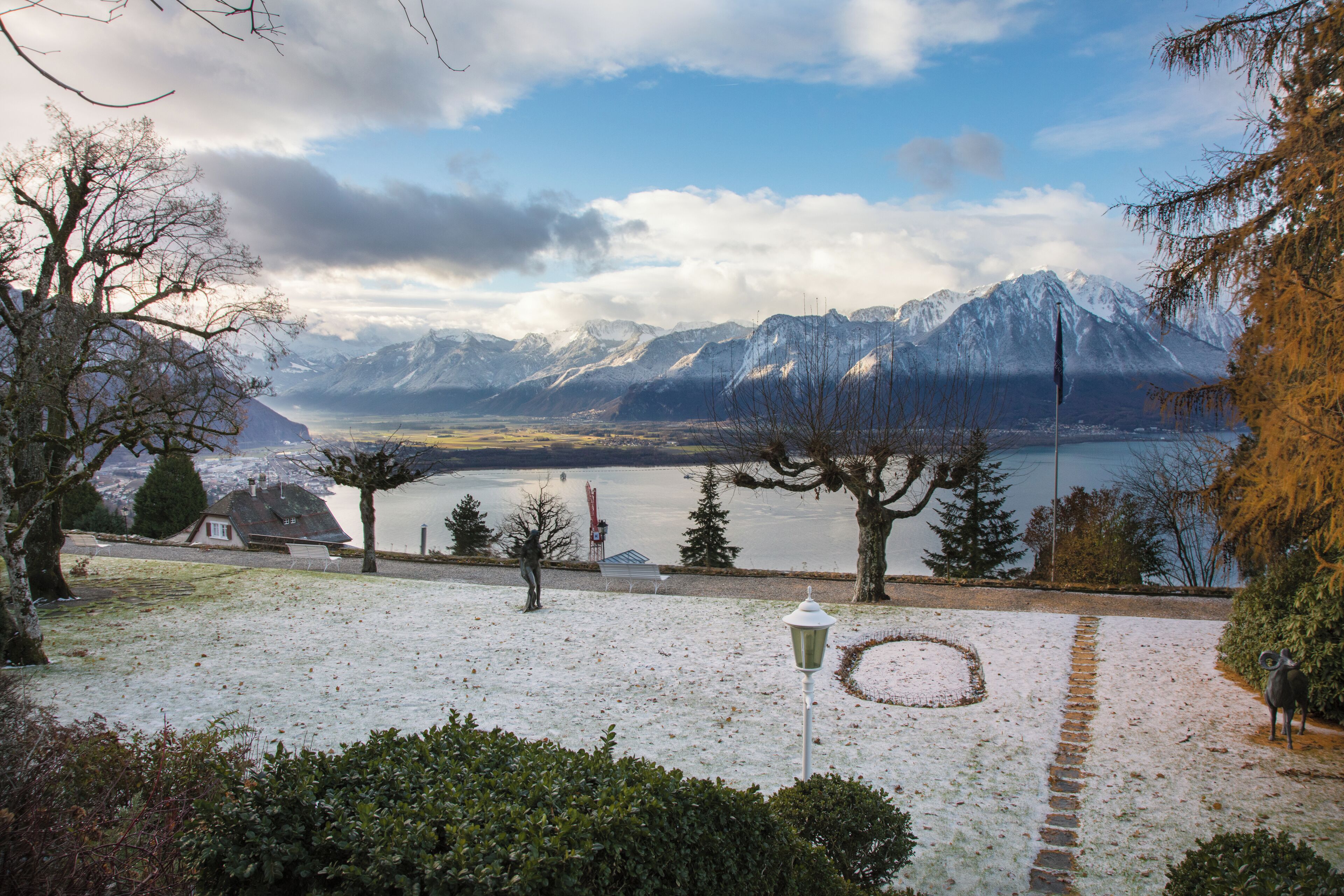 A snowy landscape with a lake, mountains, and a pathway leading to a house.