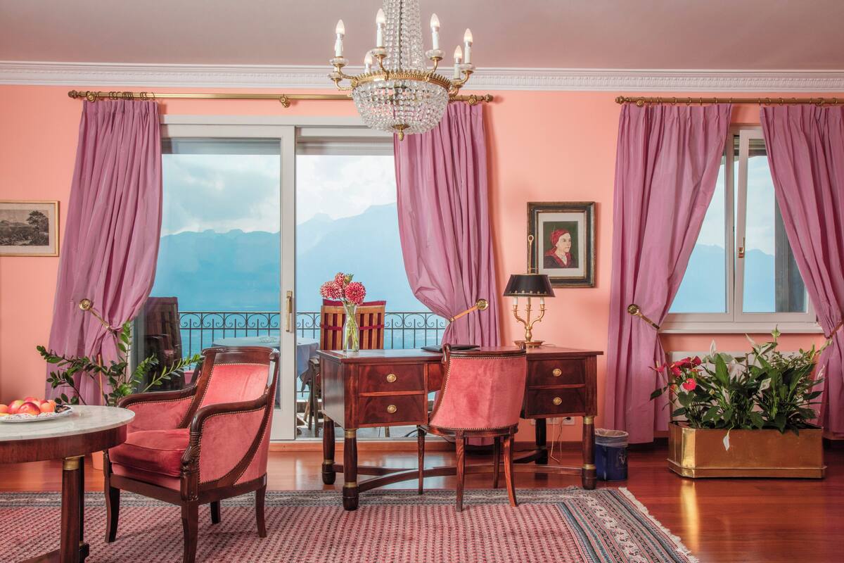 A room with a wooden desk, two chairs, a chandelier, a potted plant, and a view of mountains through a large window.