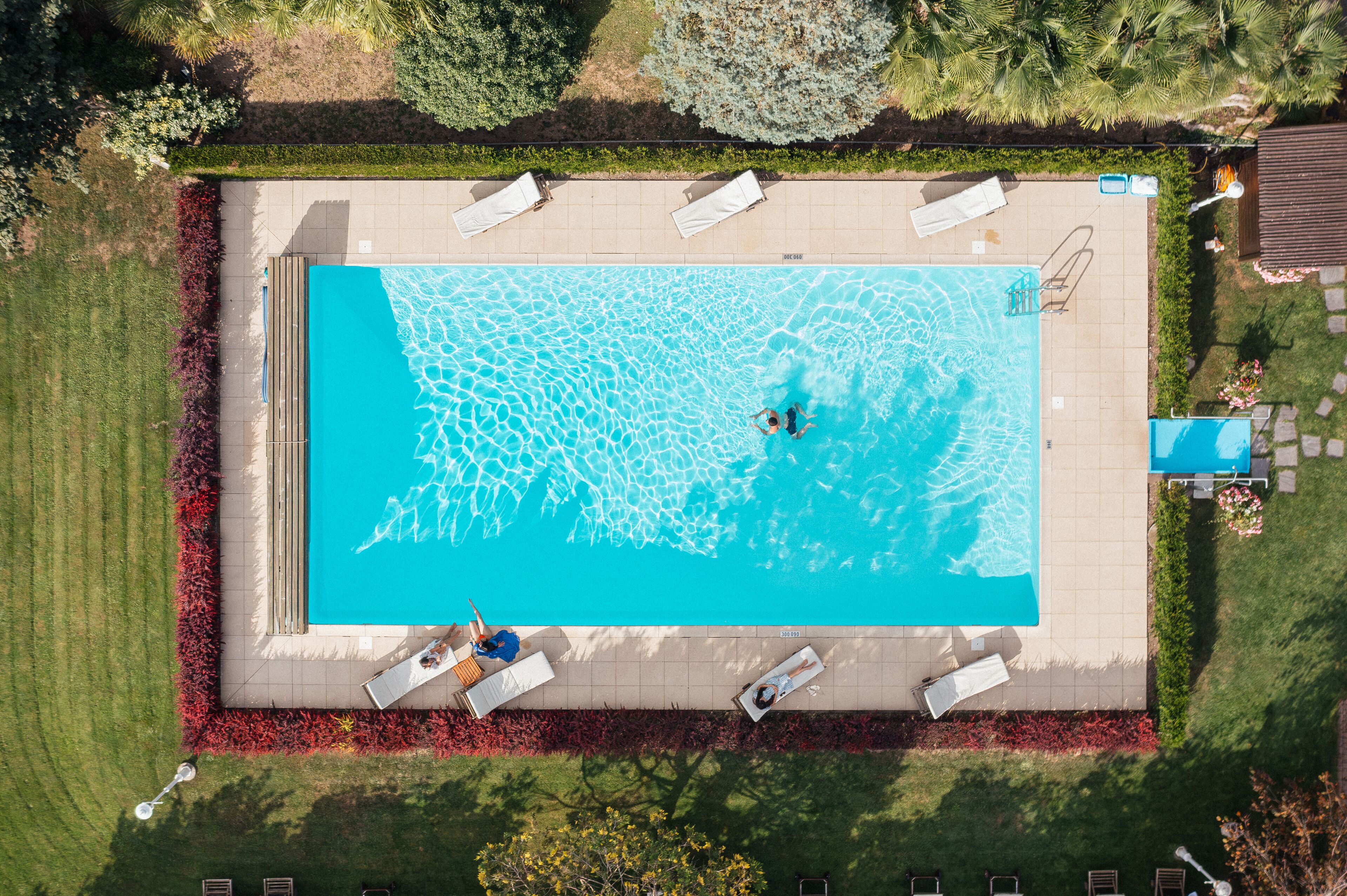 Vista aérea de una piscina rectangular con solárium empedrado, rodeada de un césped cuidado y una fila de sillones blancos.