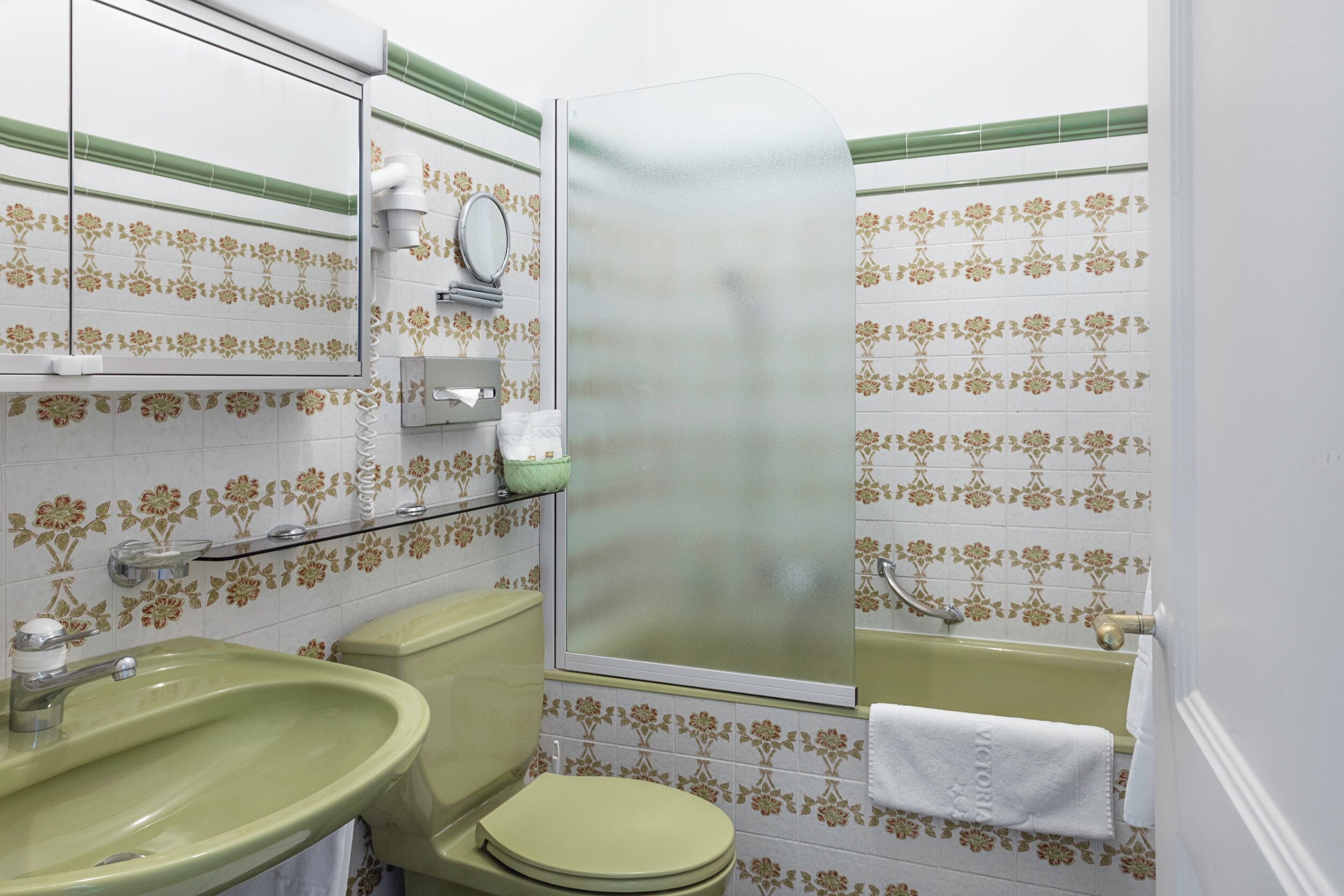 A bathroom with a green sink, toilet, and bathtub, a mirror, and floral-patterned tiles.