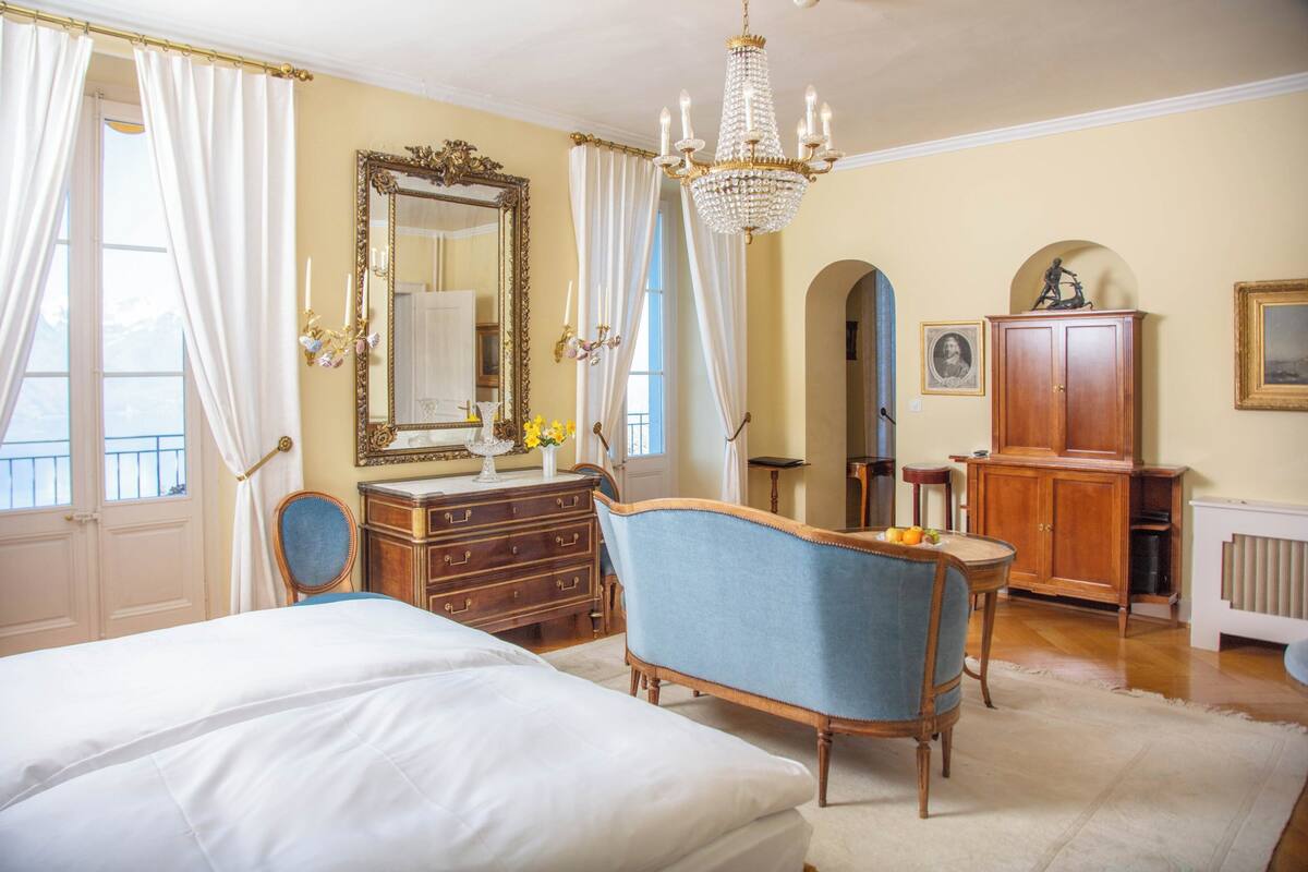 A bedroom with a large bed, a vanity with a mirror, a dresser, a blue armchair, and a chandelier.