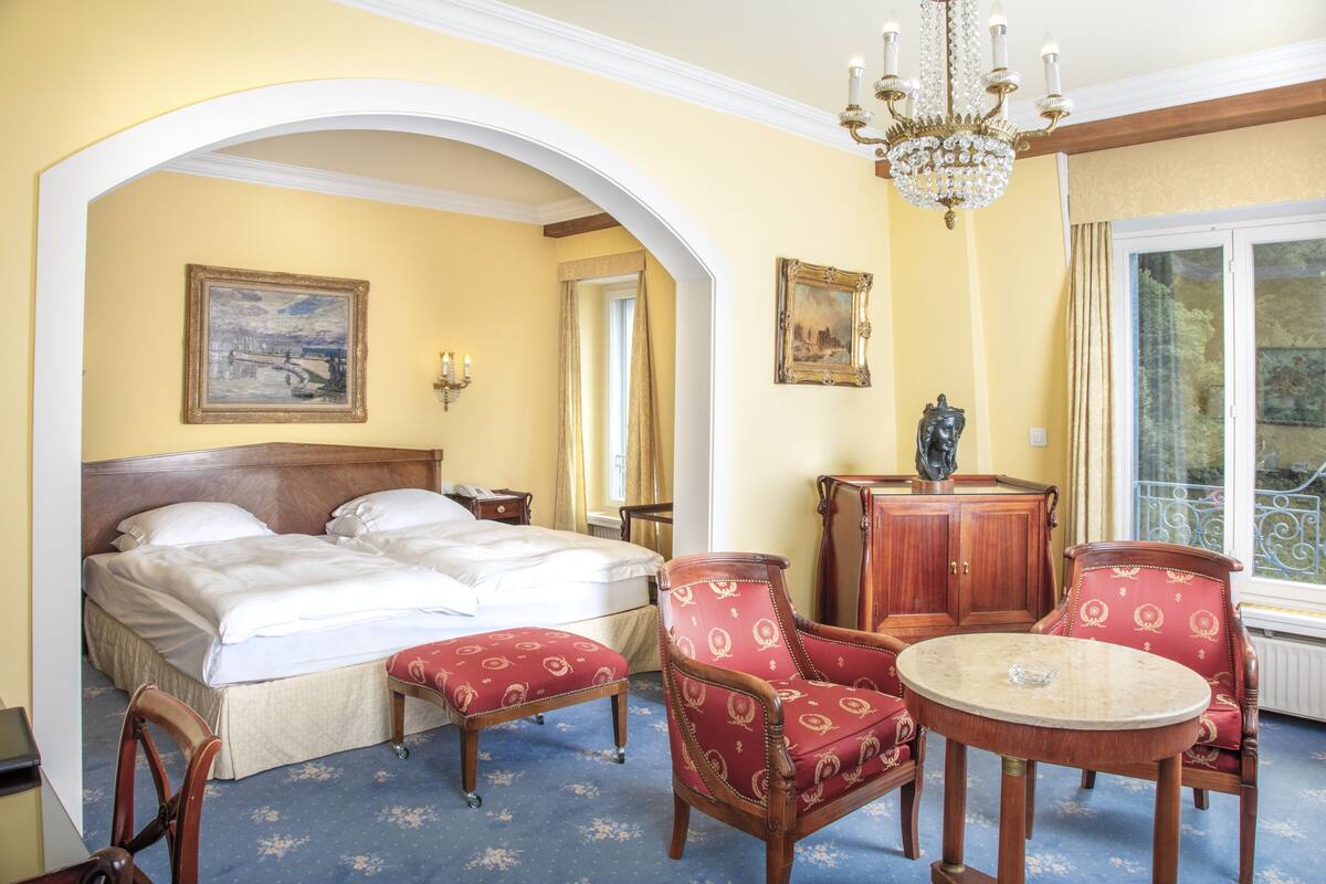 A hotel room with a bed, two red upholstered chairs, a wooden side table, and a chandelier.
